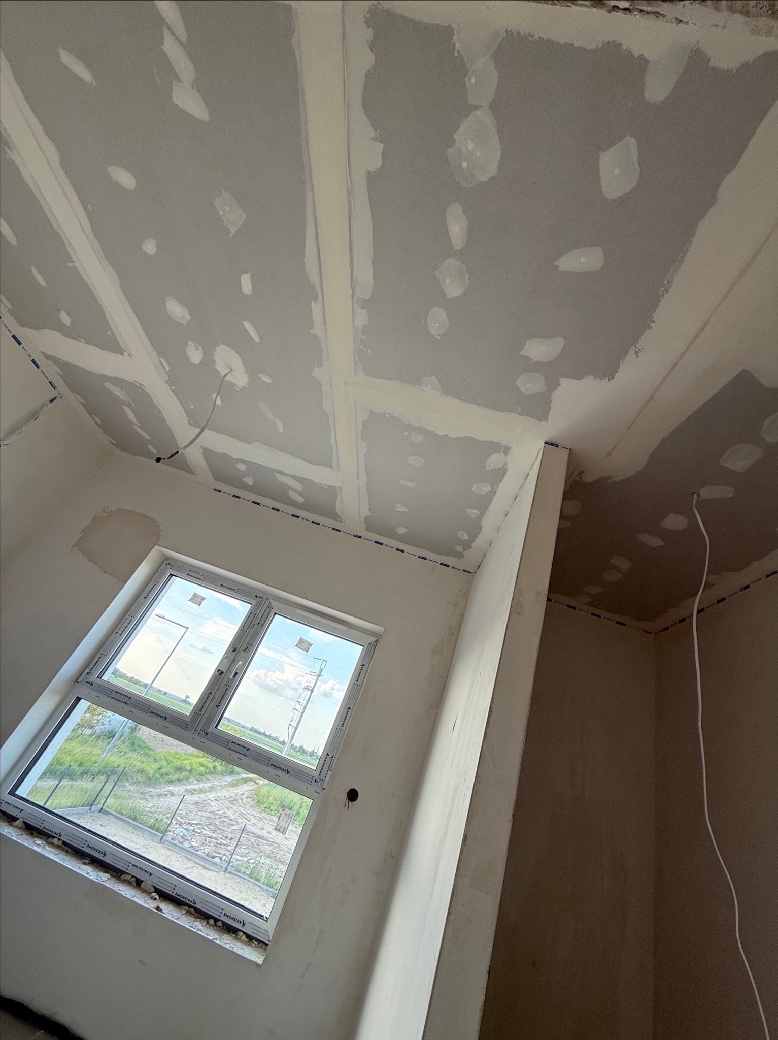 Sufit z wykończonymi łączeniami płyt gipsowych, widok z dołu, okno z widokiem na zewnątrz, przewody elektryczne. Prace wykończeniowe w surowym wnętrzu.