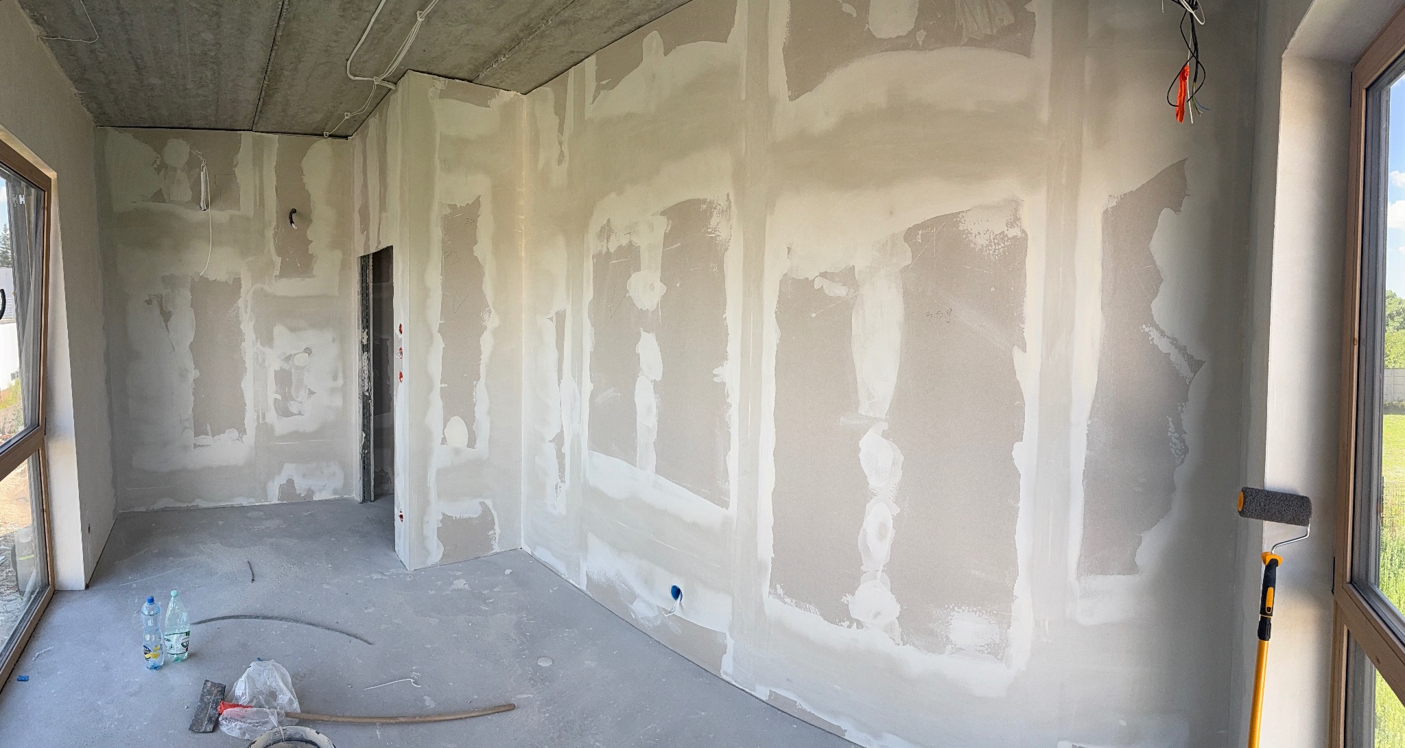 Wnętrze w trakcie prac wykończeniowych: szpachlowane ściany z widocznymi łączeniami płyt, surowy betonowy sufit, narzędzia malarskie i butelki z wodą na podłodze.