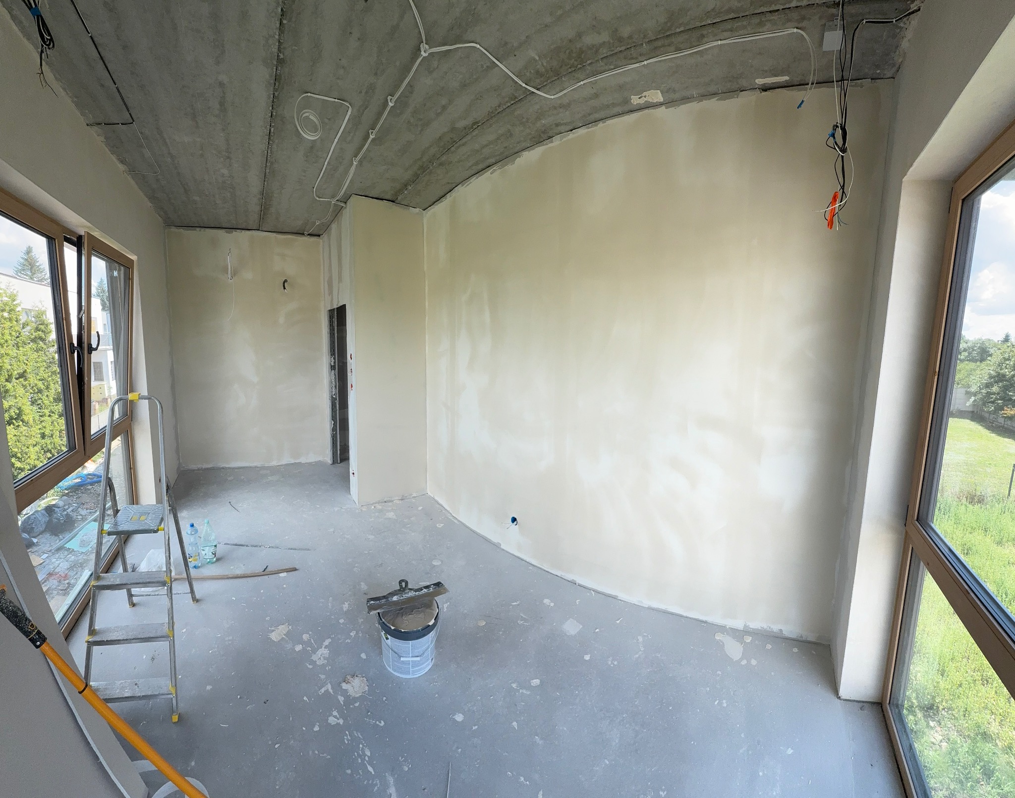 Wnętrze w trakcie prac wykończeniowych: ściany po szpachlowaniu, surowy betonowy sufit z instalacją elektryczną, okna z widokiem na zieleń. Widoczny sprzęt malarski.