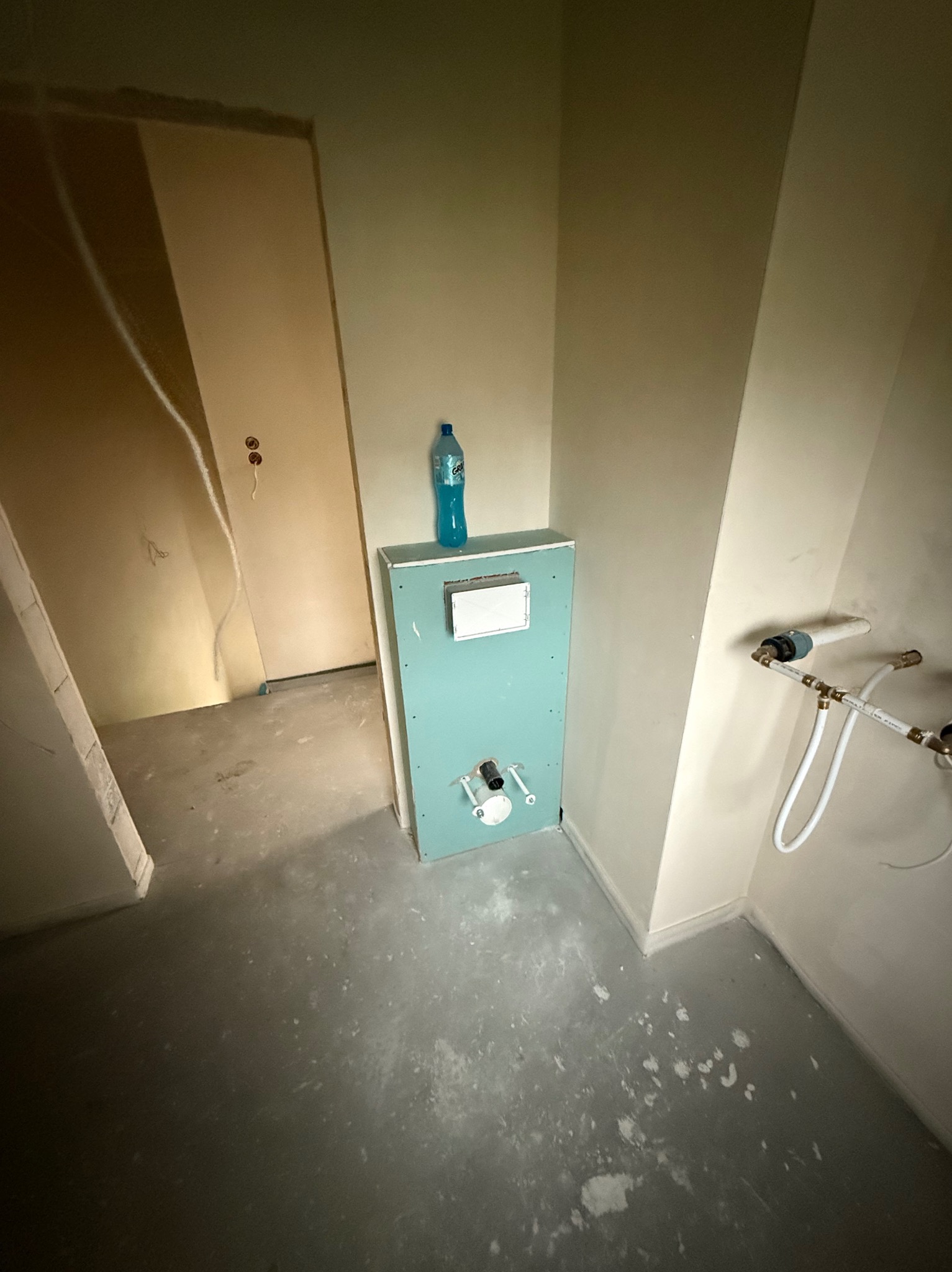 Montaż stelaża podtynkowego z płyt karton-gips w łazience, widoczne rury instalacji wodnej i butelka wody na obudowie. Szare ściany w surowym stanie.