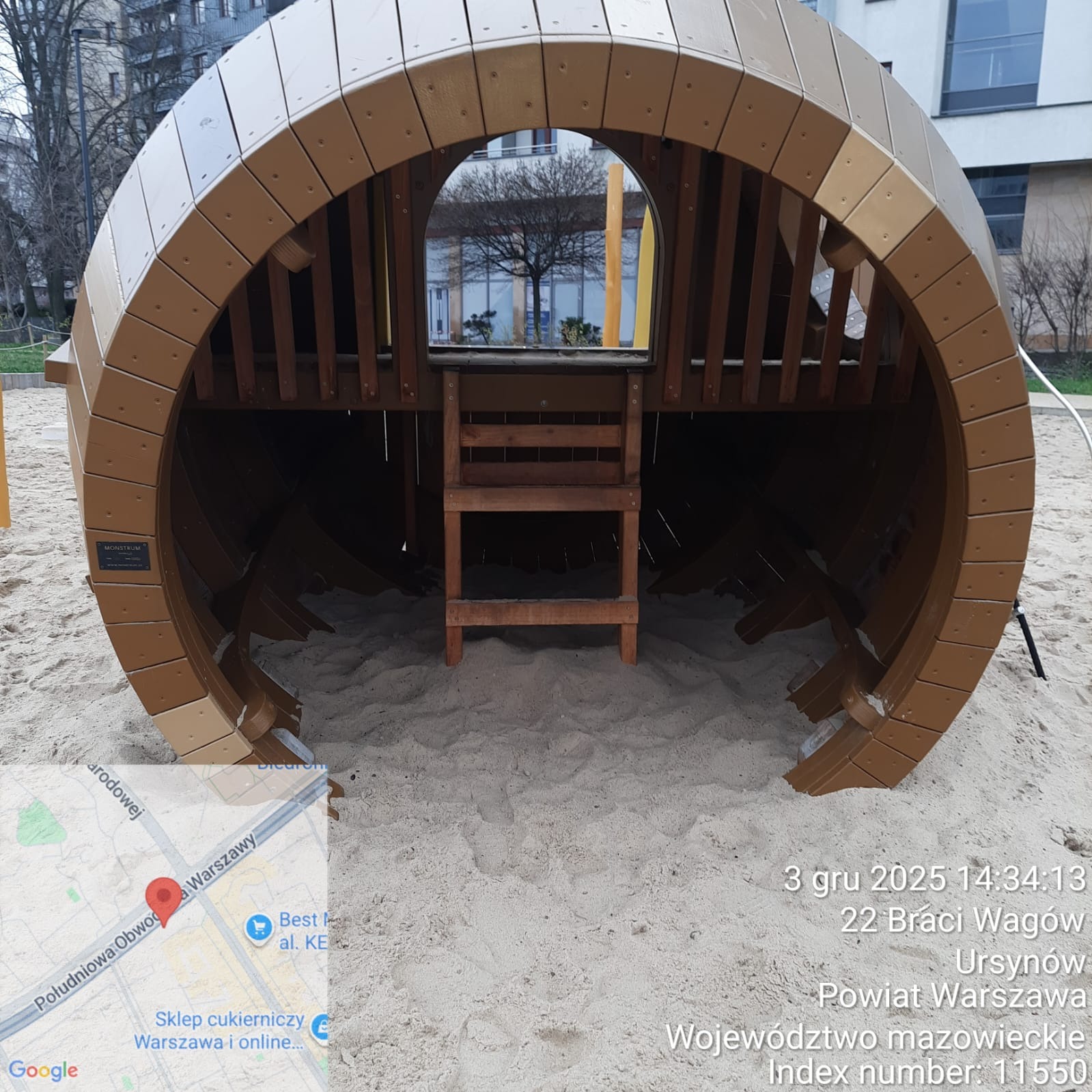 Drewniana konstrukcja w kształcie beczki z drabinką na piaskowym placu zabaw. Widok z przodu, neutralne oświetlenie dzienne. Element wyposażenia placu zabaw.