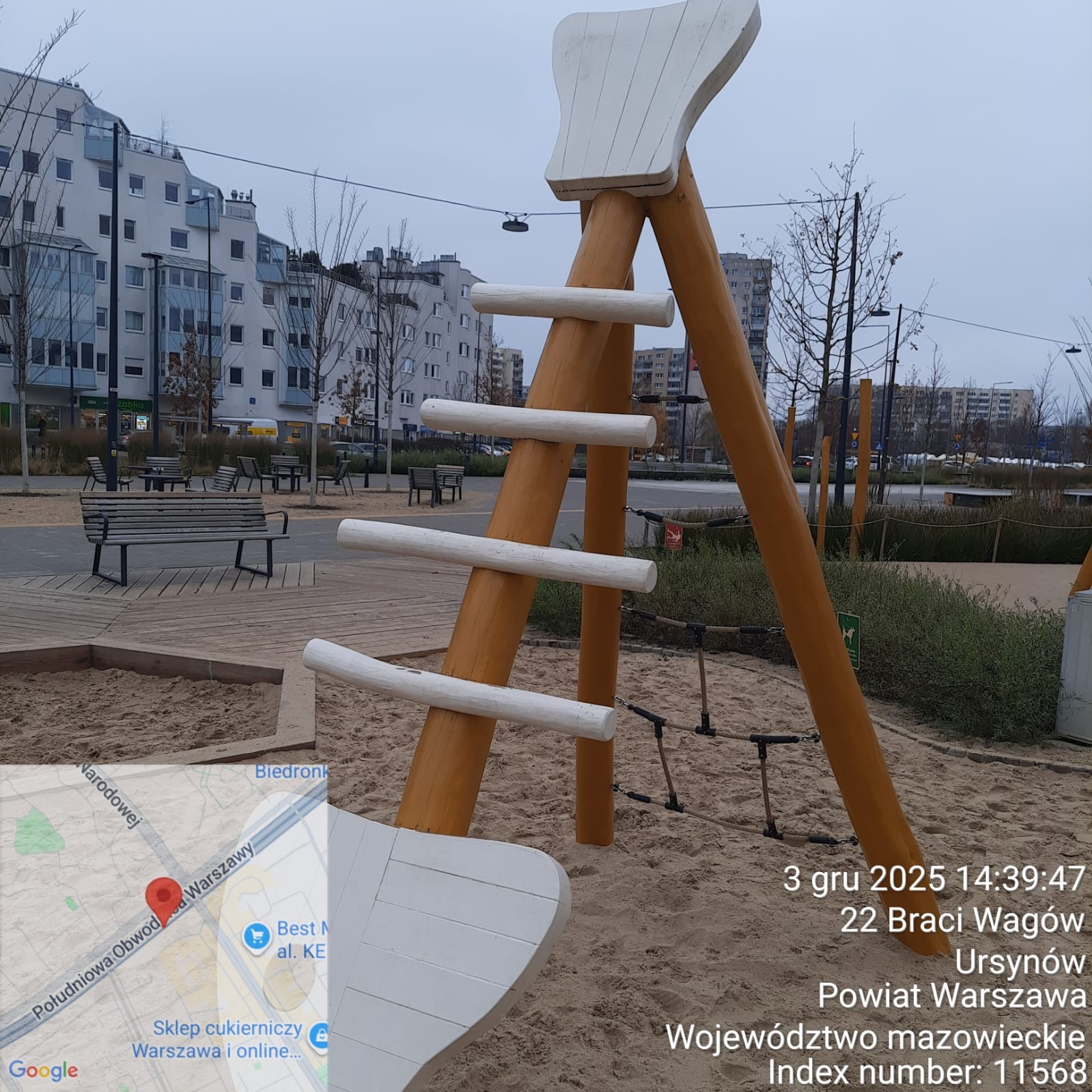 Drewniana konstrukcja w kształcie drabiny na placu zabaw, z piaskownicą i budynkami w tle. Elementy pomalowane na biało i brązowo. Widok na miejski krajobraz.