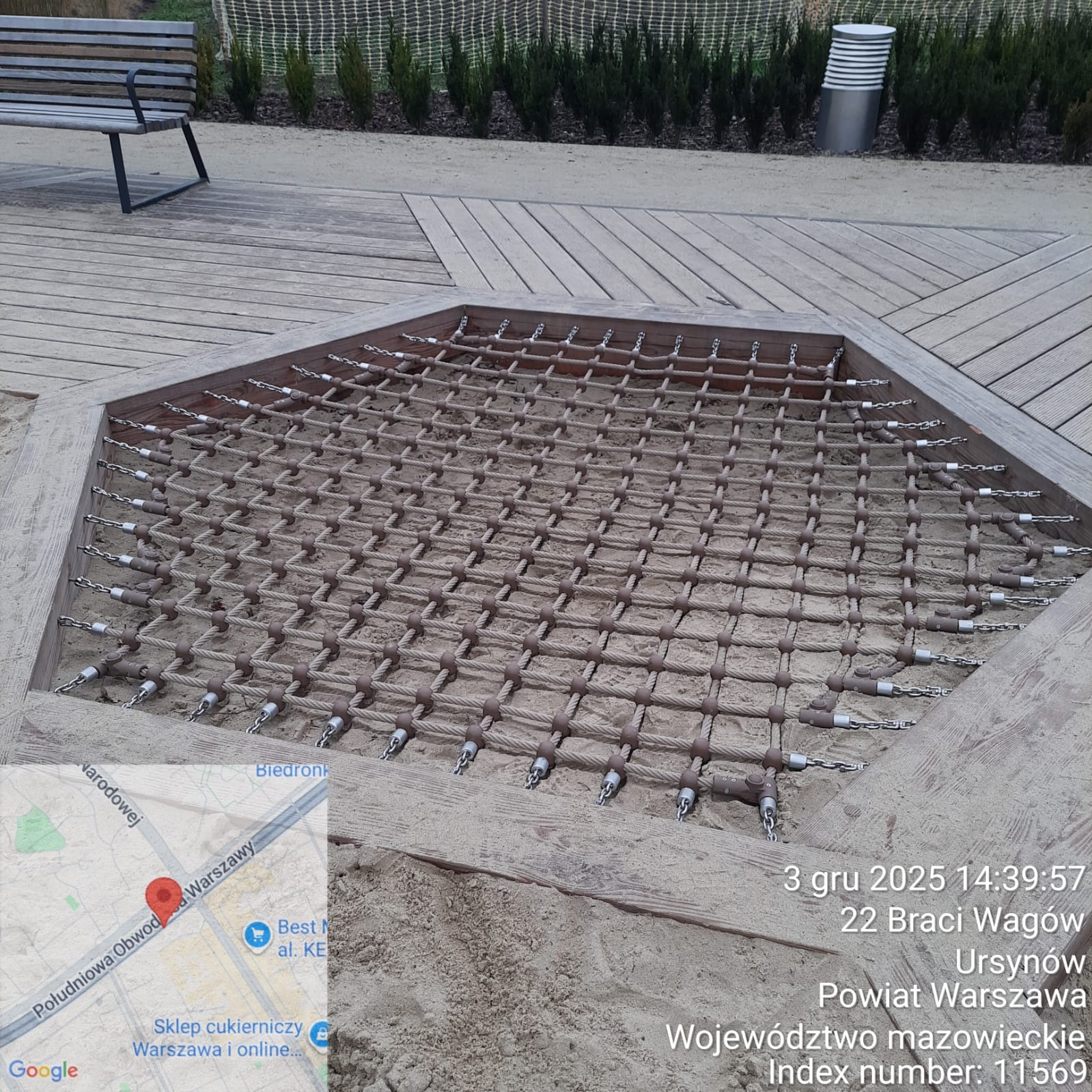 Piaskownica z siatką linową na drewnianym podeście, element placu zabaw. Widoczne łańcuchy mocujące i piasek. W tle ławka i roślinność.