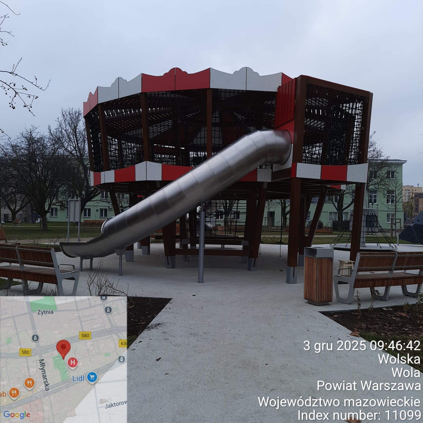 Nowoczesny plac zabaw z metalową zjeżdżalnią i konstrukcją z siatki, otoczony zielenią miejską. Elementy w kolorach czerwieni i bieli. Widoczne ławki i kosze na śmieci.