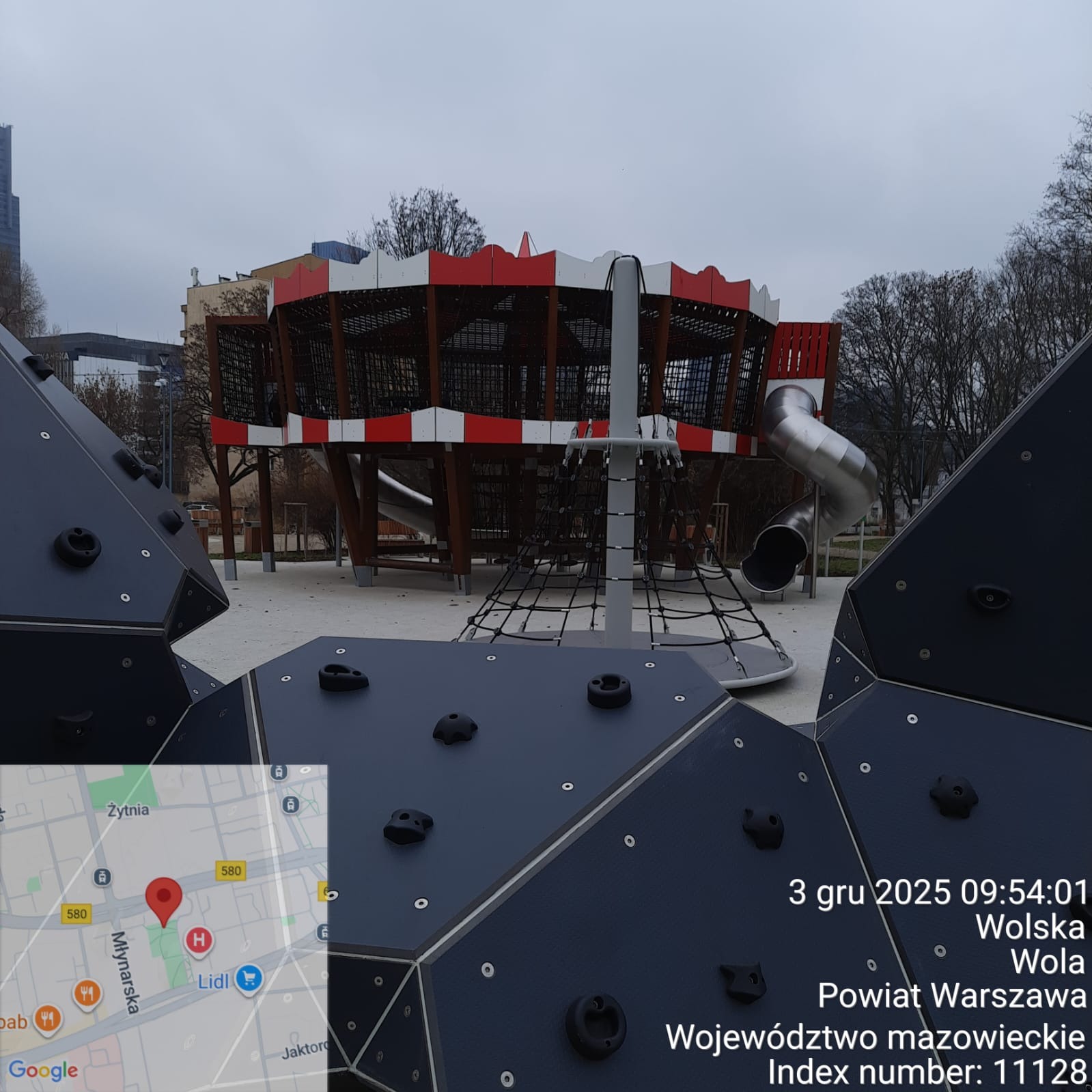 Nowoczesny plac zabaw z konstrukcją w kształcie statku, ścianką wspinaczkową i zjeżdżalnią. Elementy w kolorach czerwieni, bieli i szarości. Widok z dołu na tle zabudowy miejskiej.