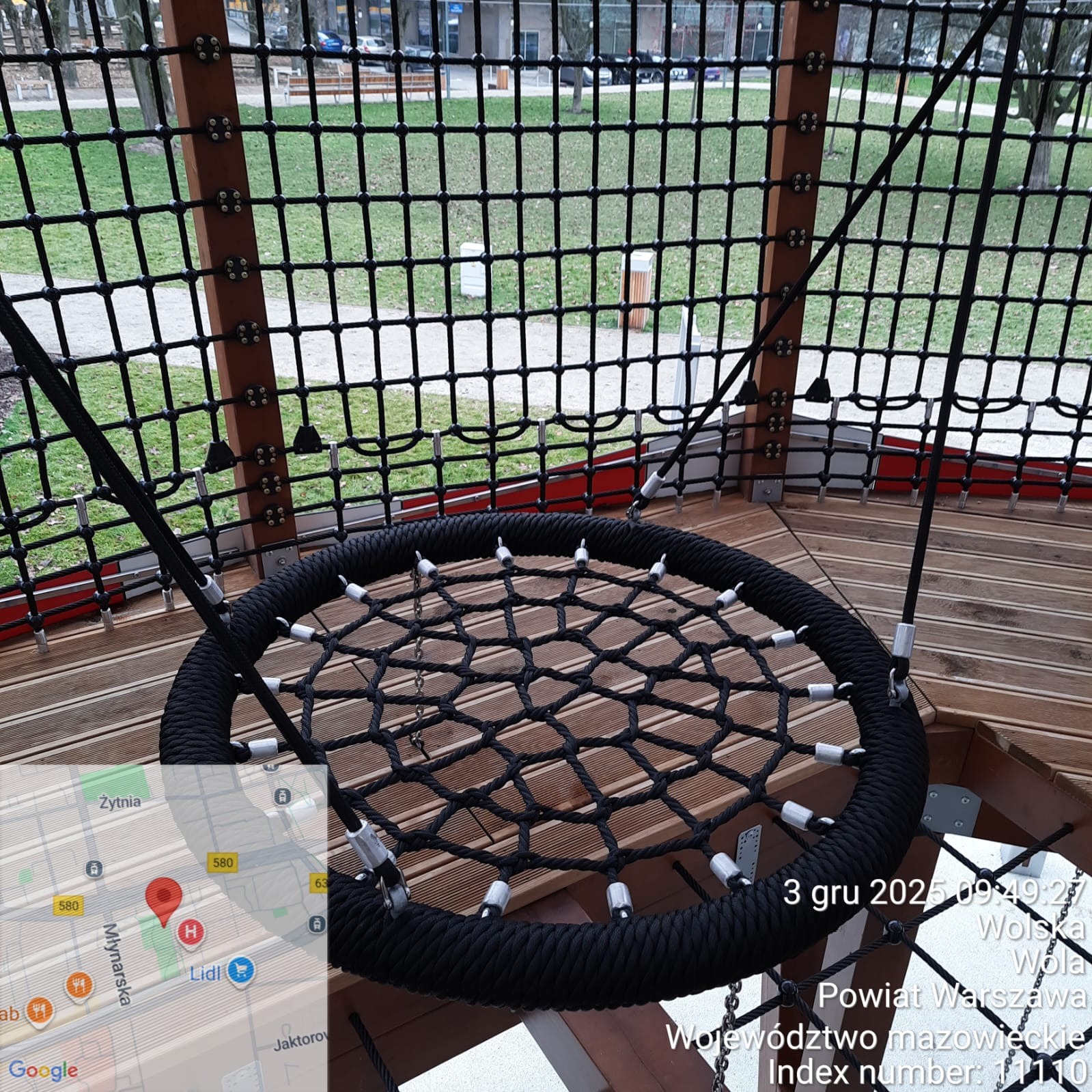 Huśtawka typu 'gniazdo' z czarnej liny na drewnianej konstrukcji placu zabaw, widok z góry, siatka zabezpieczająca w tle. Widoczny fragment mapy z lokalizacją.