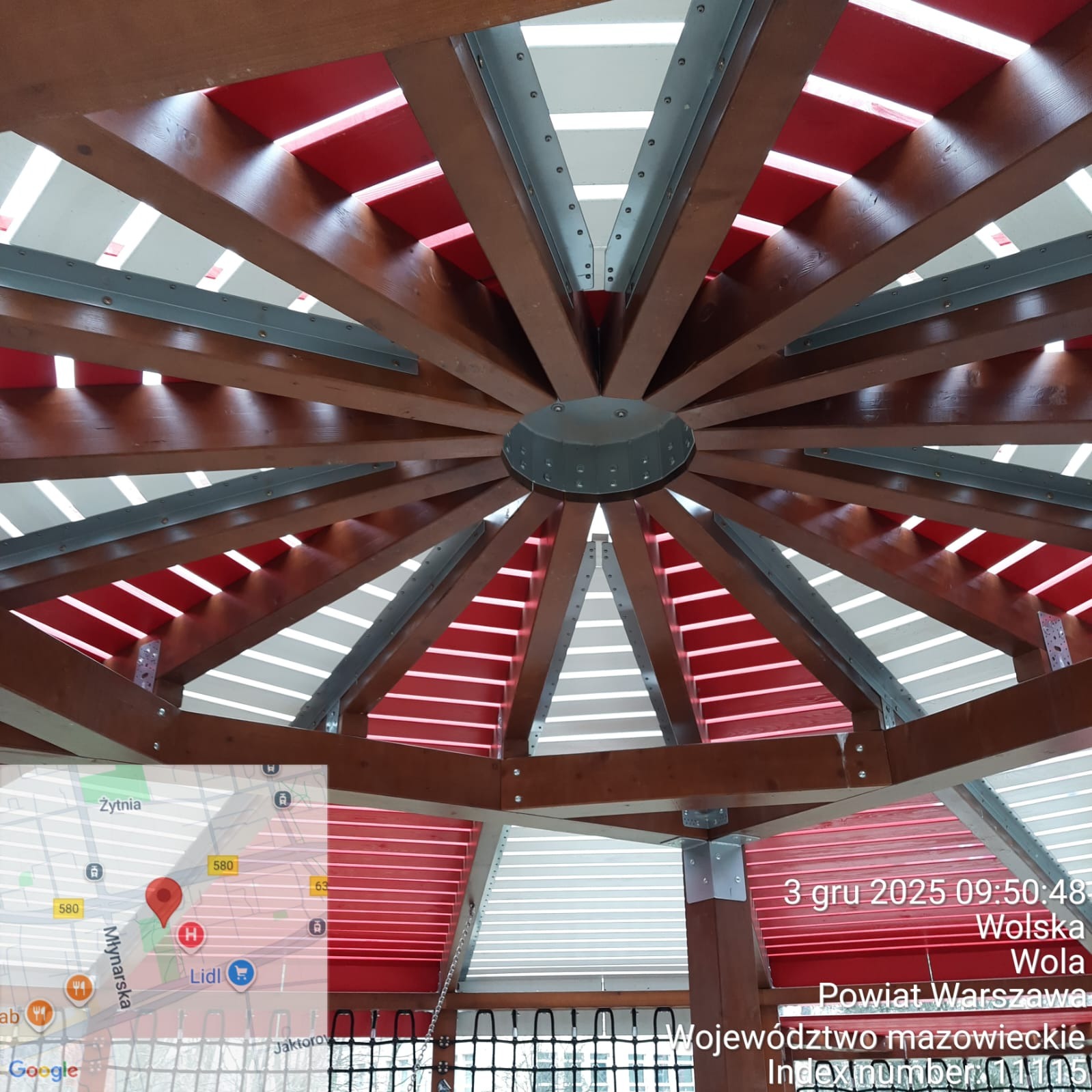 Widok z dołu na drewnianą konstrukcję z czerwonymi i białymi elementami, prawdopodobnie zadaszenie placu zabaw. Metalowe łączniki. Częściowo widoczna mapa i dane lokalizacyjne.