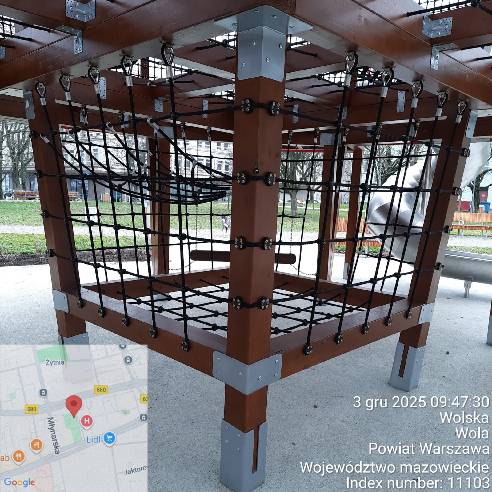 Drewniana konstrukcja linowa placu zabaw z metalowymi łącznikami, widok z dołu, na tle zieleni parku miejskiego. Elementy konstrukcji częściowo poza kadrem.