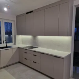 Kremowa kuchnia z czarnym zlewem i płytą grzewczą. Górne szafki podświetlone, biały blat i marmurowe wykończenie ściany. Widoczne okno i niedokończone oświetlenie sufitowe.