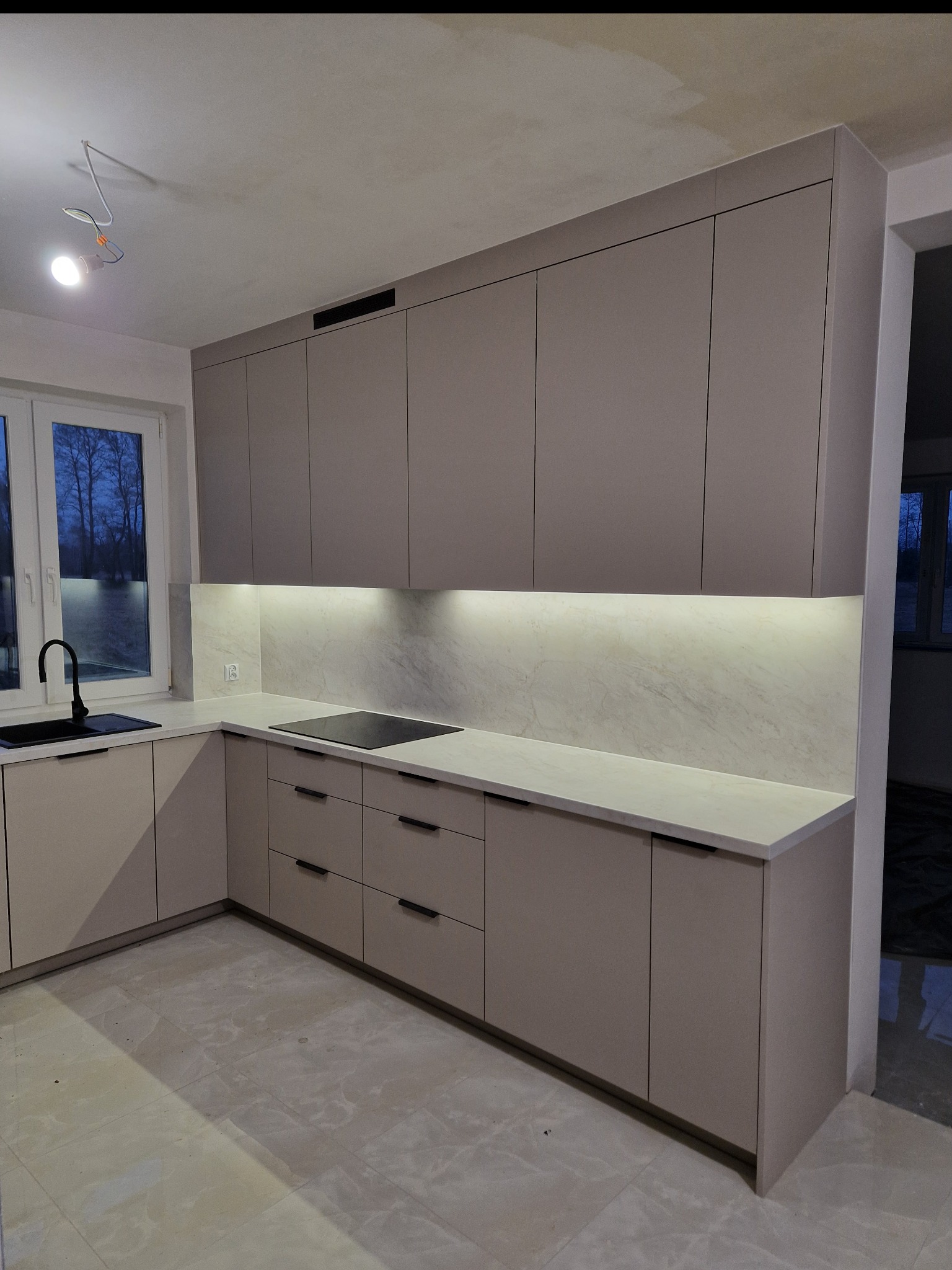 Kremowa kuchnia z czarnym zlewem i płytą grzewczą. Górne szafki podświetlone, biały blat i marmurowe wykończenie ściany. Widoczne okno i niedokończone oświetlenie sufitowe.