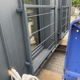 SPAWPRO - Szary, modułowy budynek z oknami i metalową balustradą na platformie transportowej. Widoczne kable instalacji i fragment niebieskiej maszyny.