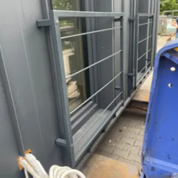 Szary, modułowy budynek z oknami i metalową balustradą na platformie transportowej. Widoczne kable instalacji i fragment niebieskiej maszyny.