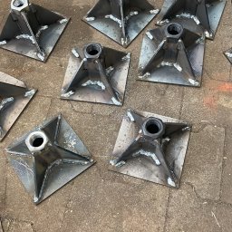 SPAWPRO - Metalowe podstawy z nakrętkami, spawane na kwadratowych płytach, ułożone na betonowej posadzce. Widoczne ślady spawania i niedoskonałości obróbki.