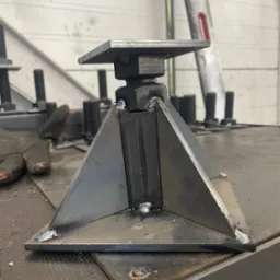 Metalowa konstrukcja spawana z płaskownikiem i kwadratową podstawą, widoczne spawy, obok leżą rękawice robocze, na tle ściany z blachy trapezowej.