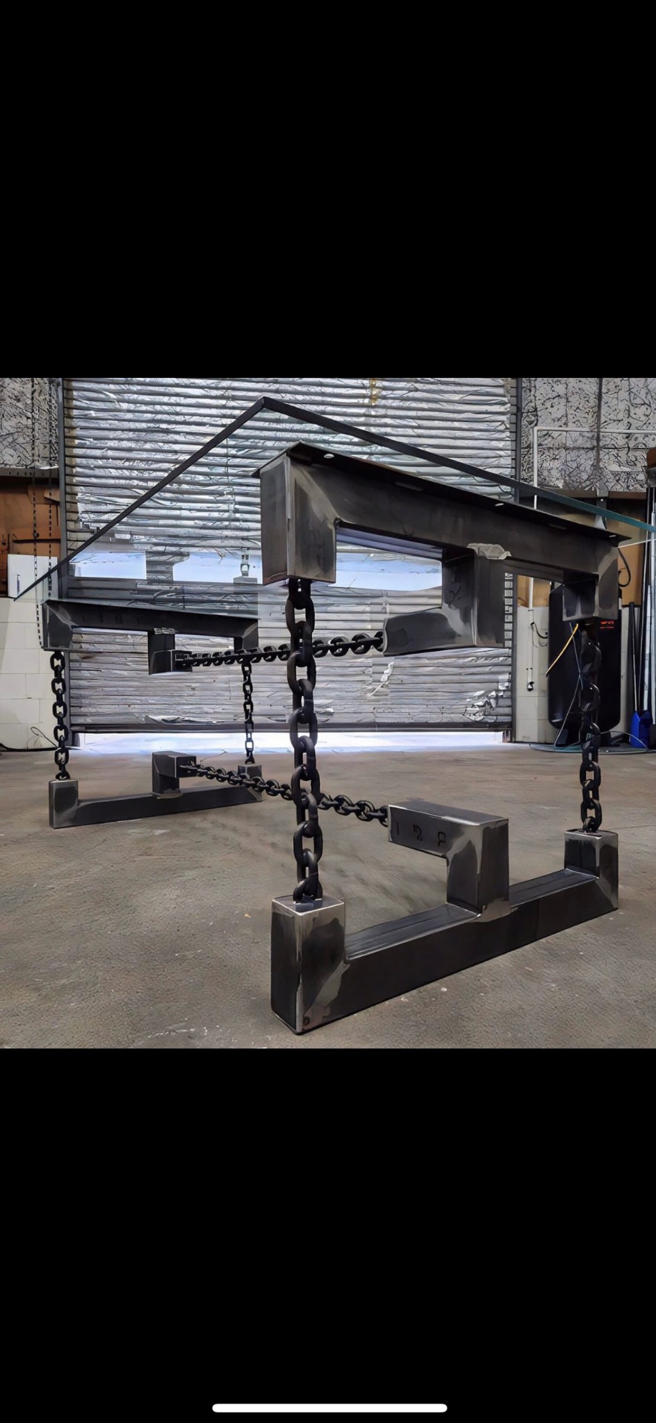 Metalowa konstrukcja przypominająca stół z elementami łańcucha, surowy industrialny styl, widoczne spawy, szara podłoga i brama garażowa w tle.