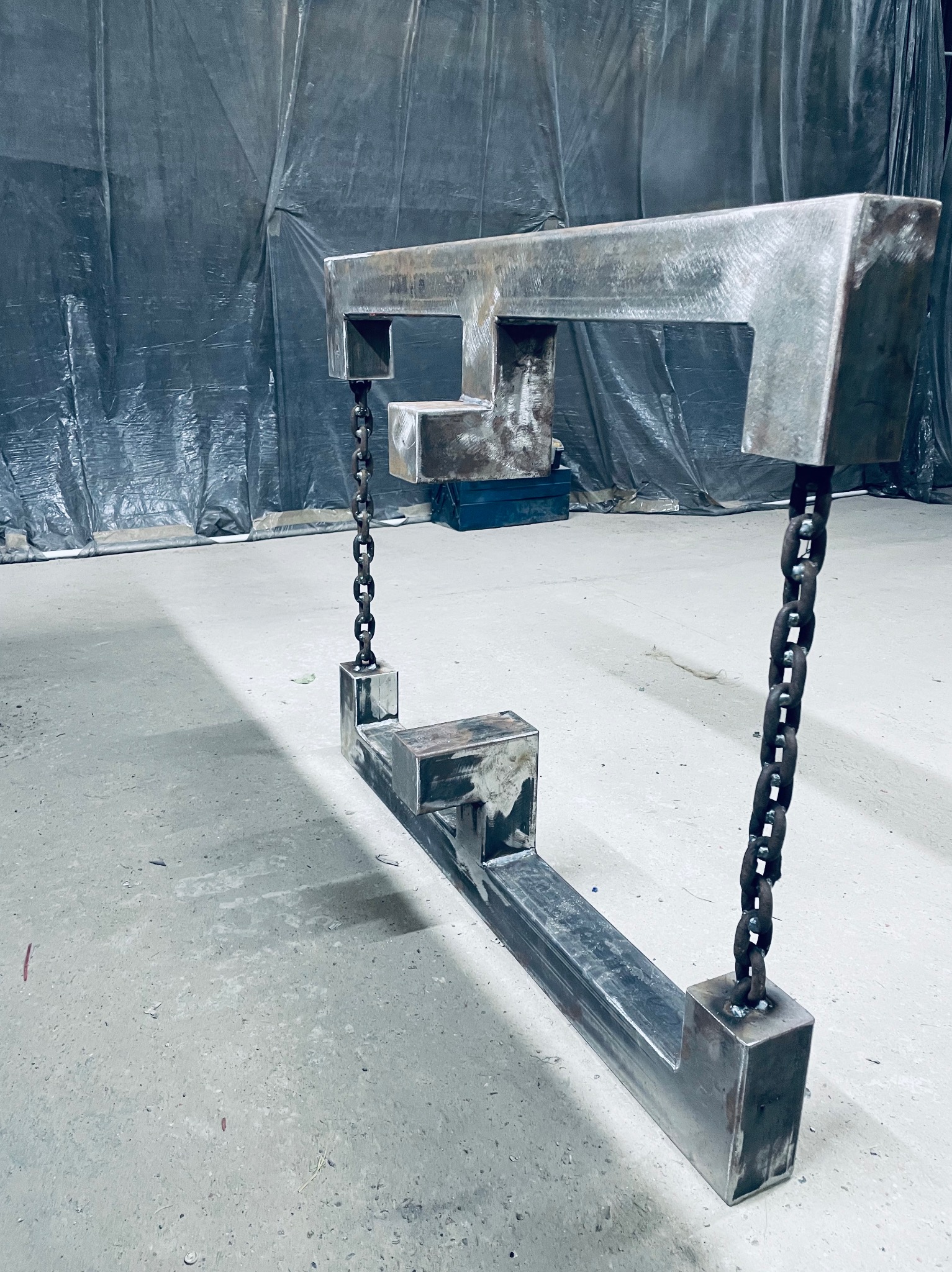 Surowa, metalowa konstrukcja spawana z łańcuchami, prawdopodobnie element większej instalacji, na tle folii ochronnej w warsztacie.