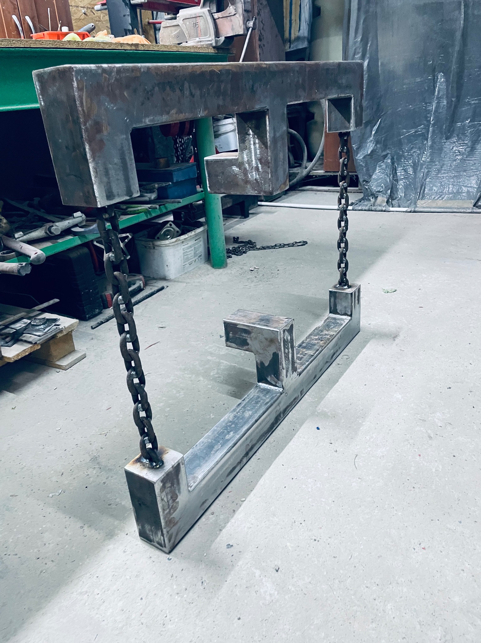 Nierównomiernie wyszlifowany, metalowy element konstrukcyjny zawieszony na łańcuchach w warsztacie. Surowy, industrialny wygląd.