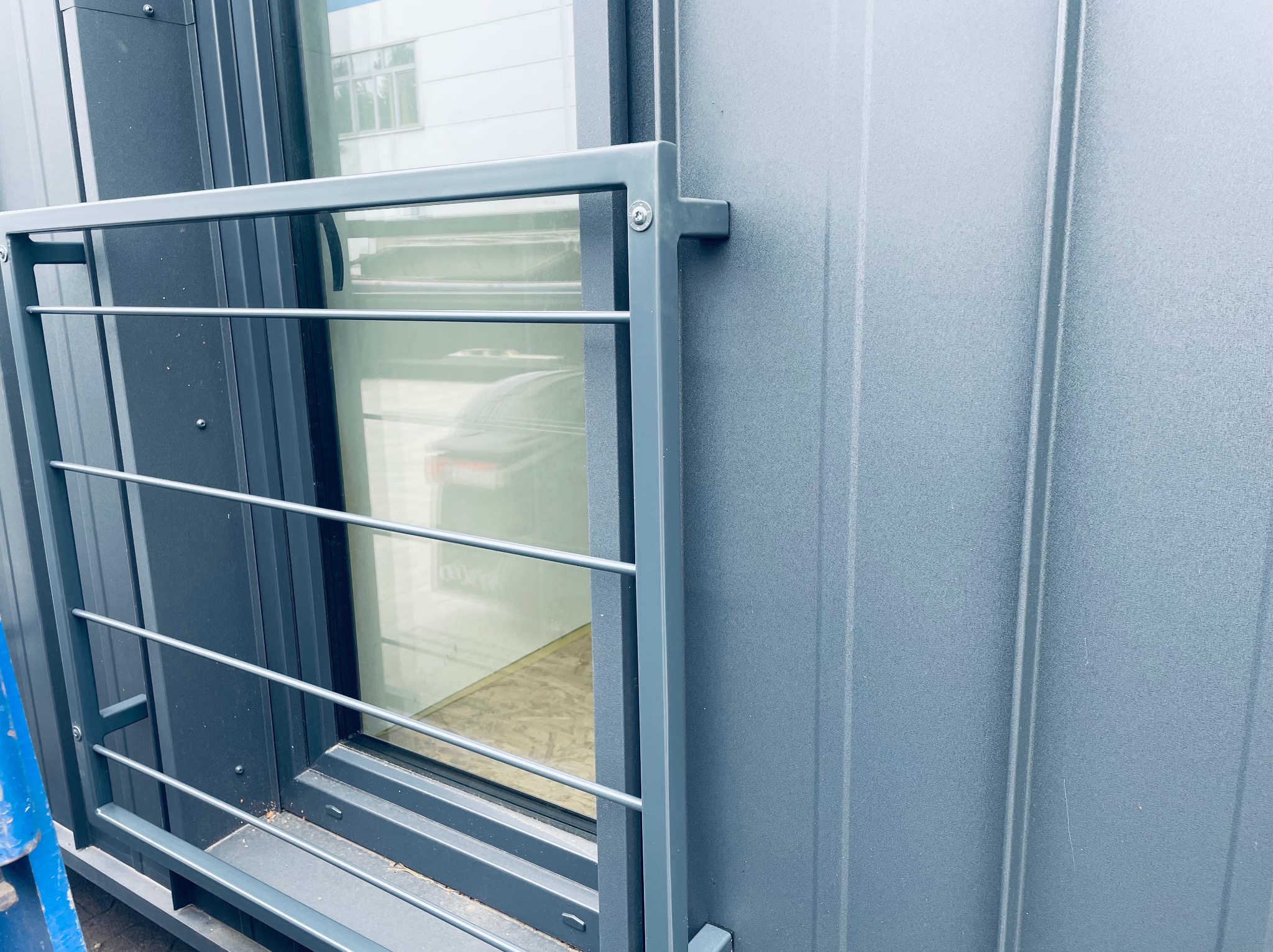 Szara, metalowa balustrada balkonowa z poziomymi prętami, zamontowana przy oknie w budynku o szarej elewacji z blachy trapezowej. Widok z bliska.