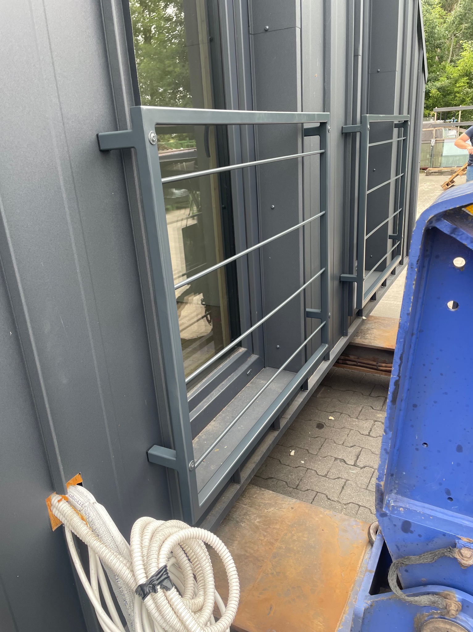 Szary, modułowy budynek z oknami i metalową balustradą na platformie transportowej. Widoczne kable instalacji i fragment niebieskiej maszyny.