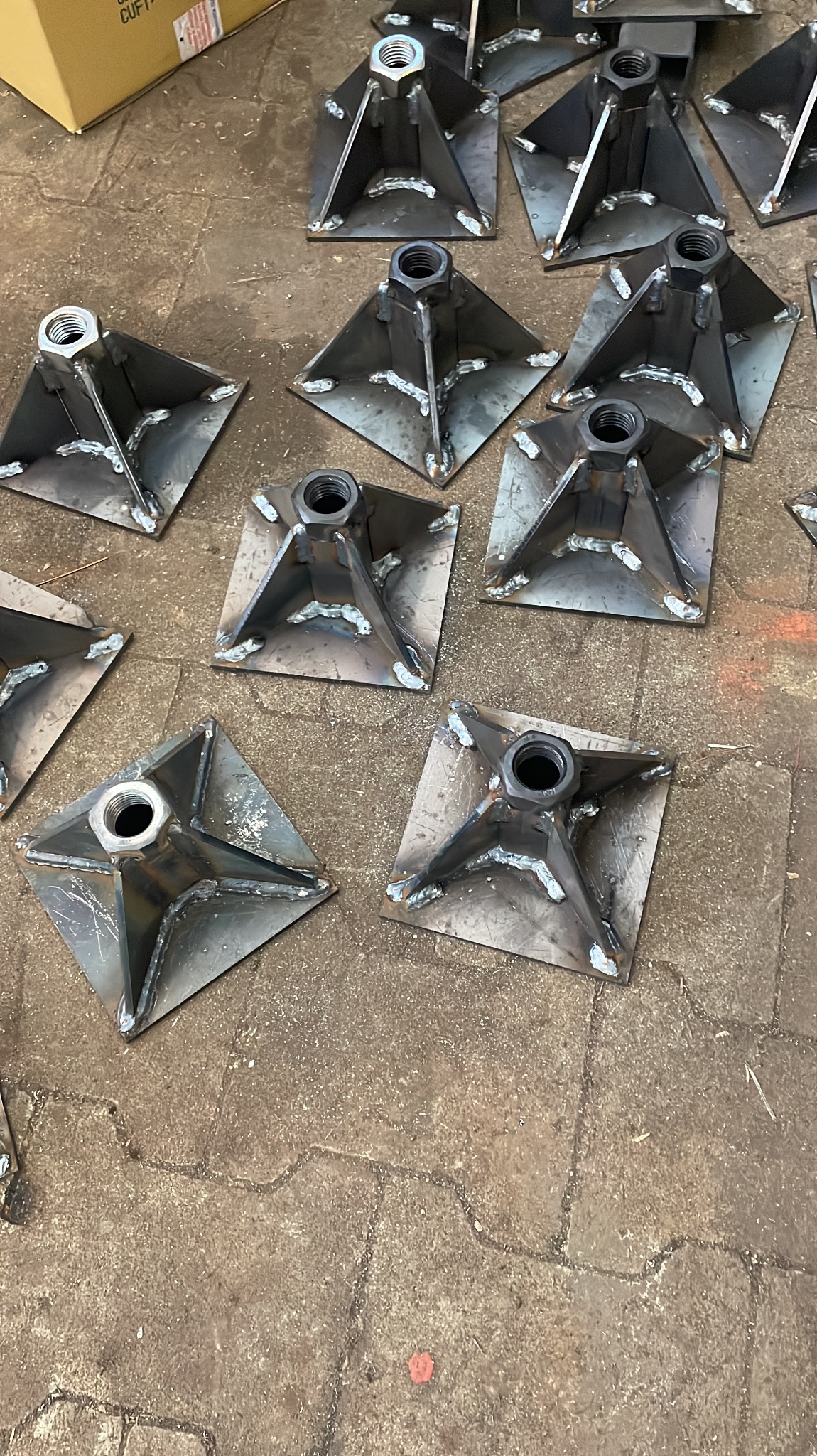 Metalowe podstawy z nakrętkami, spawane na kwadratowych płytach, ułożone na betonowej posadzce. Widoczne ślady spawania i niedoskonałości obróbki.