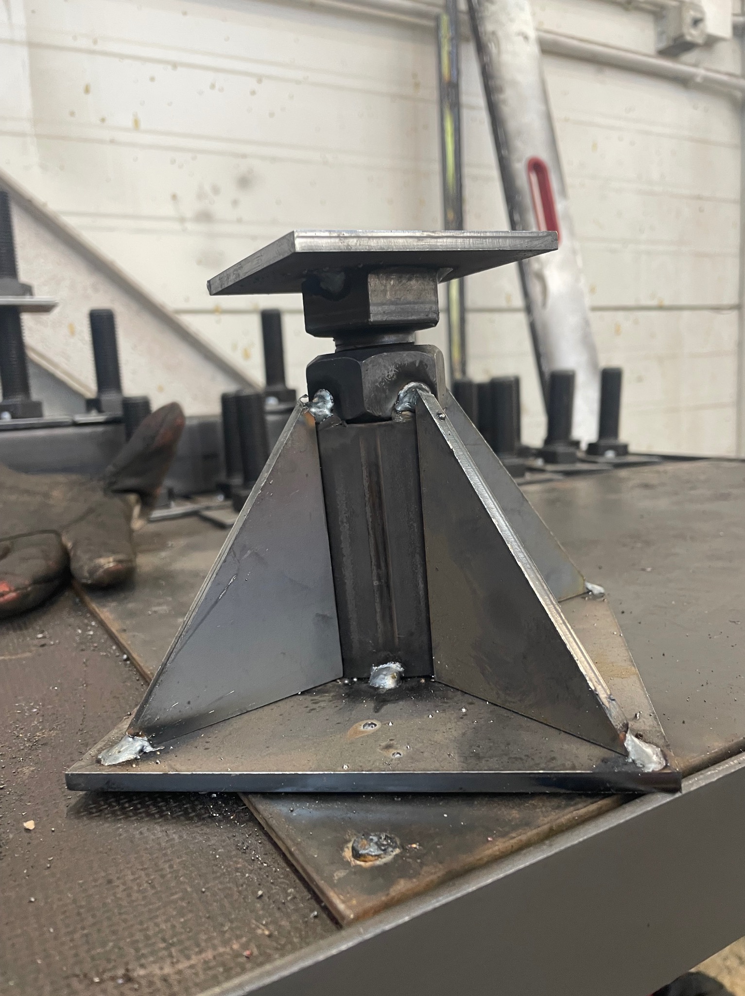 Metalowa konstrukcja spawana z płaskownikiem i kwadratową podstawą, widoczne spawy, obok leżą rękawice robocze, na tle ściany z blachy trapezowej.
