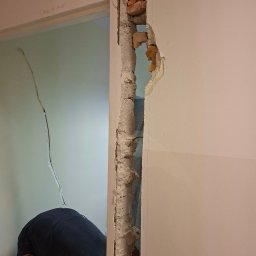 Artur Bozek - Częściowo wyburzona ściana z cegły, widoczny fragment muru i instalacji. W tle sylwetka osoby w trakcie pracy, oświetlona przestrzeń sąsiedniego pomieszczenia.