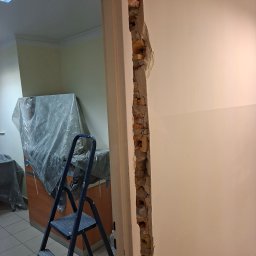 Artur Bozek - Fragment ściany po wyburzeniu z widocznymi cegłami, drabina w tle. Prace remontowe w pomieszczeniu z zabezpieczonymi meblami folią.