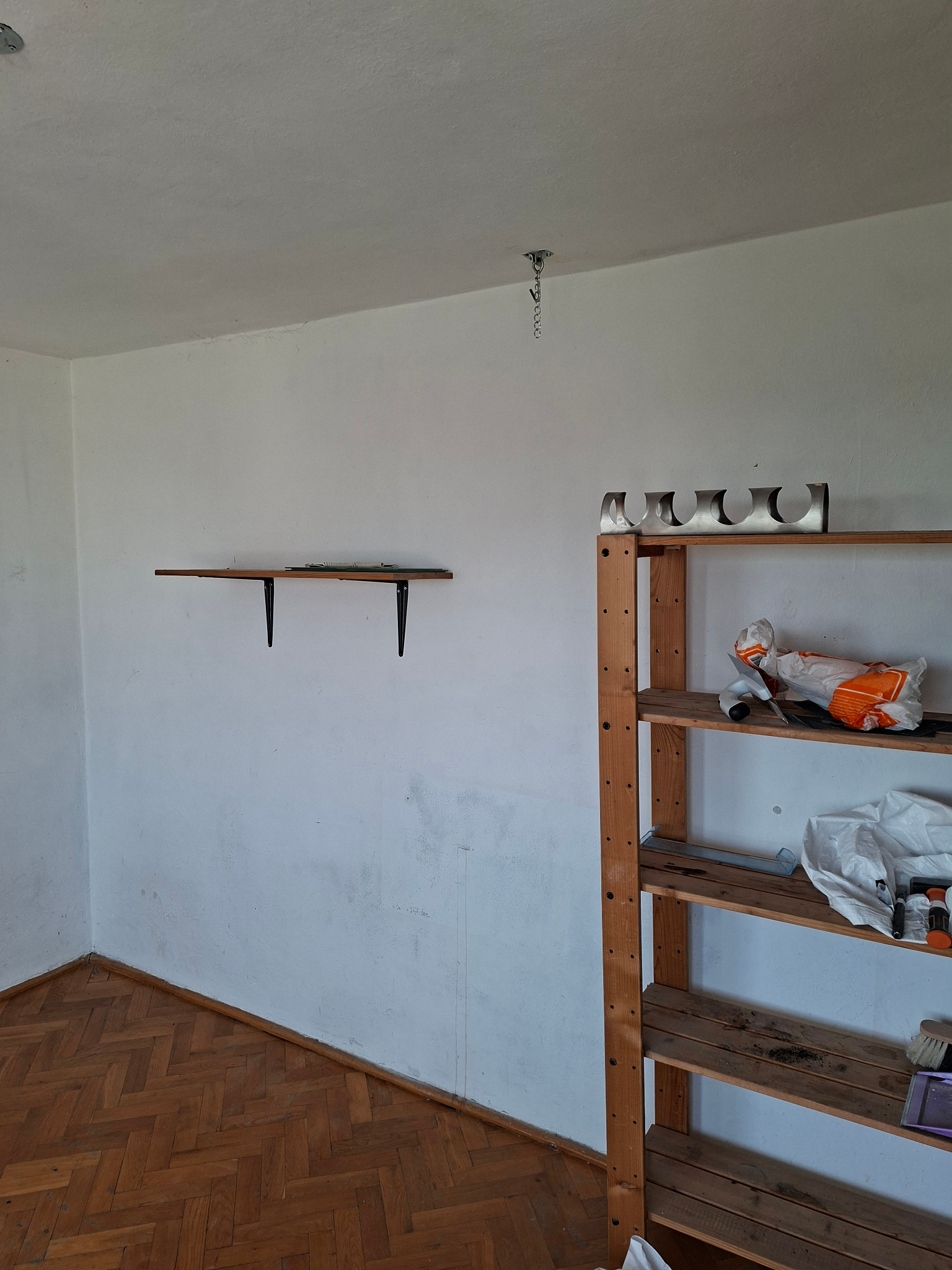 Pusty pokój z drewnianą podłogą w jodełkę, białe ściany z półką i regałem. Widoczne ślady użytkowania i przygotowania do remontu. Wisząca lampa na suficie.