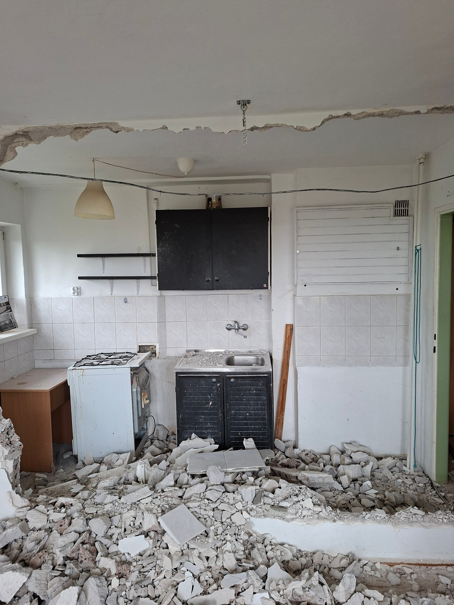 Zdemolowana kuchnia z gruzem na podłodze po wyburzeniu ściany. Widoczne fragmenty instalacji, stare meble i płytki. Ślady po usuniętych elementach na ścianach i suficie.