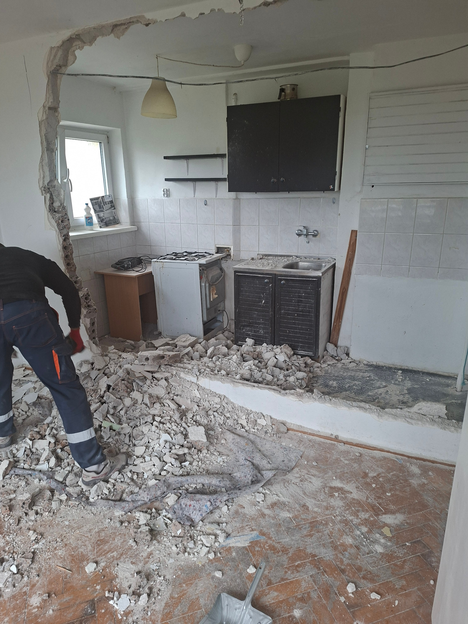 Demontaż ściany w kuchni: gruz na podłodze, widoczne instalacje. Pracownik w odzieży roboczej z narzędziem. Fragment okna i wyposażenia kuchennego w tle.