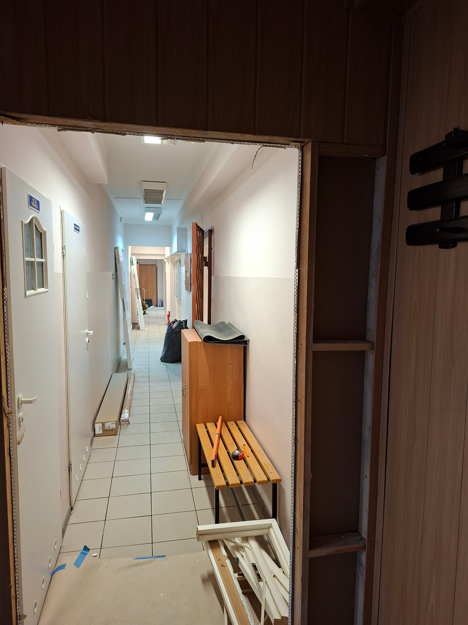 Korytarz w trakcie demontażu ścianki działowej, widoczne narzędzia i elementy konstrukcyjne. Fragment ściany z boazerią po prawej stronie kadru.