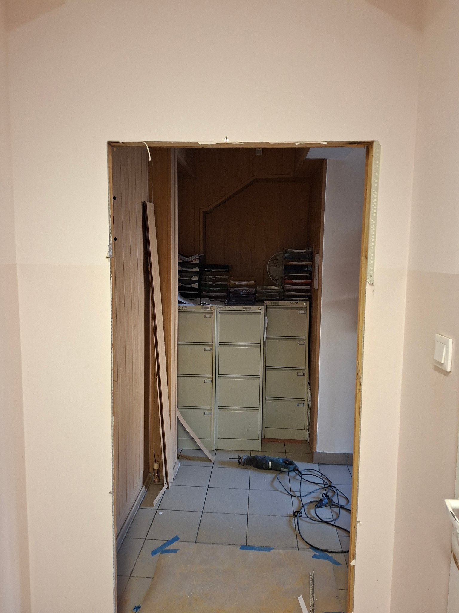 Otwór po wyburzonej ścianie działowej, widoczne narzędzia i fragmenty ścianki. W tle szafy biurowe z dokumentami. Prace wykończeniowe w trakcie realizacji.