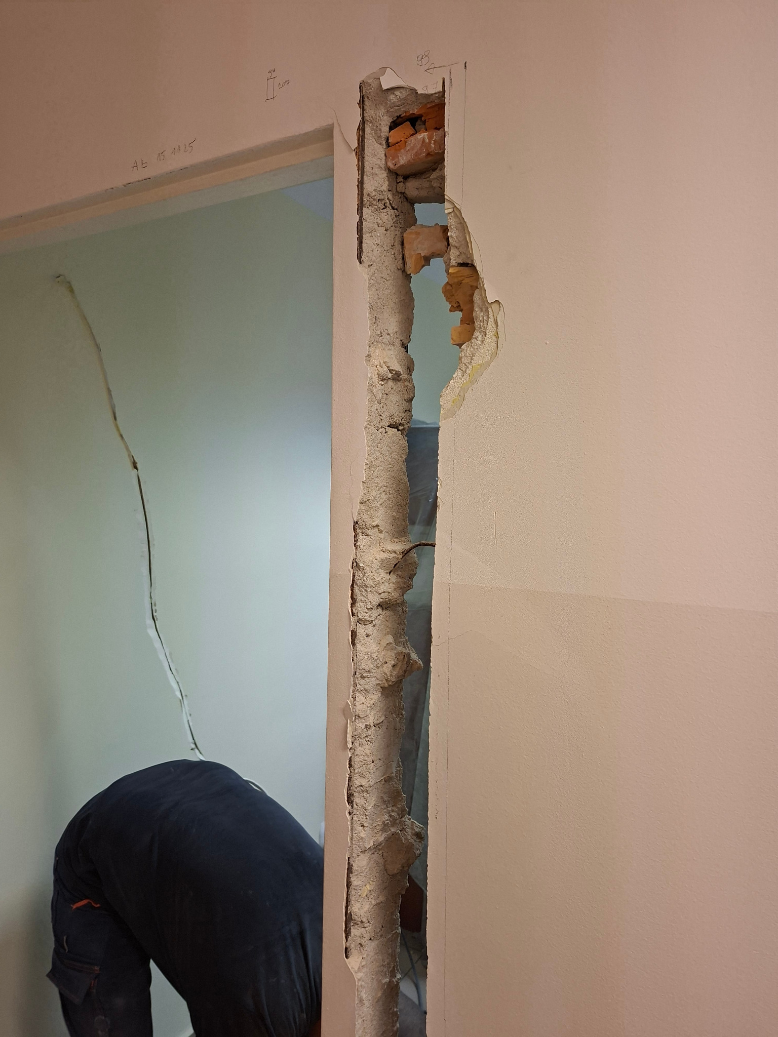 Częściowo wyburzona ściana z cegły, widoczny fragment muru i instalacji. W tle sylwetka osoby w trakcie pracy, oświetlona przestrzeń sąsiedniego pomieszczenia.