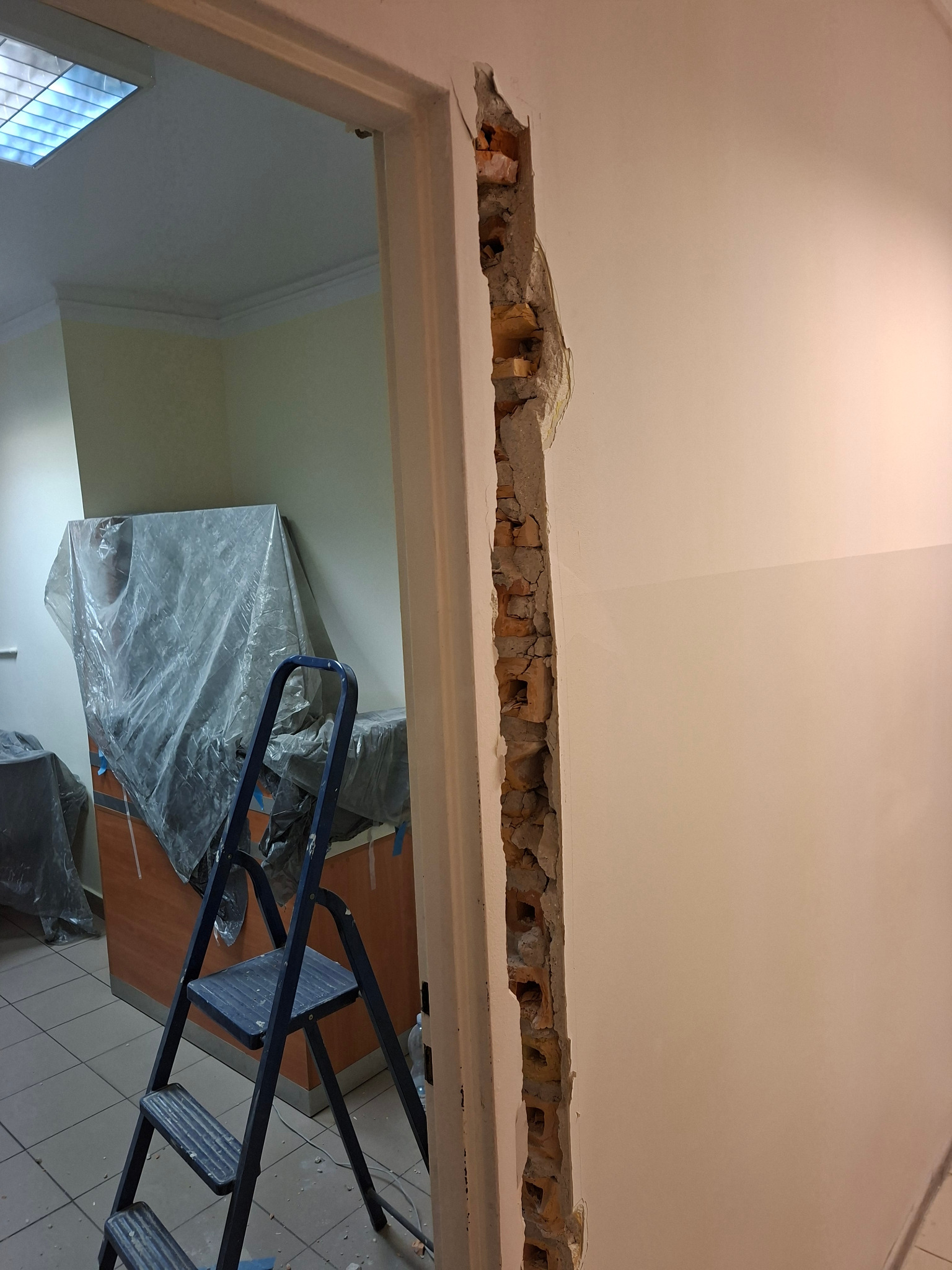 Fragment ściany po wyburzeniu z widocznymi cegłami, drabina w tle. Prace remontowe w pomieszczeniu z zabezpieczonymi meblami folią.