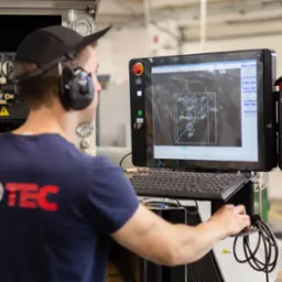 Operator CNC w słuchawkach nadzoruje proces obróbki na maszynie, obserwując schemat na monitorze. Widoczna klawiatura i panel sterowania.