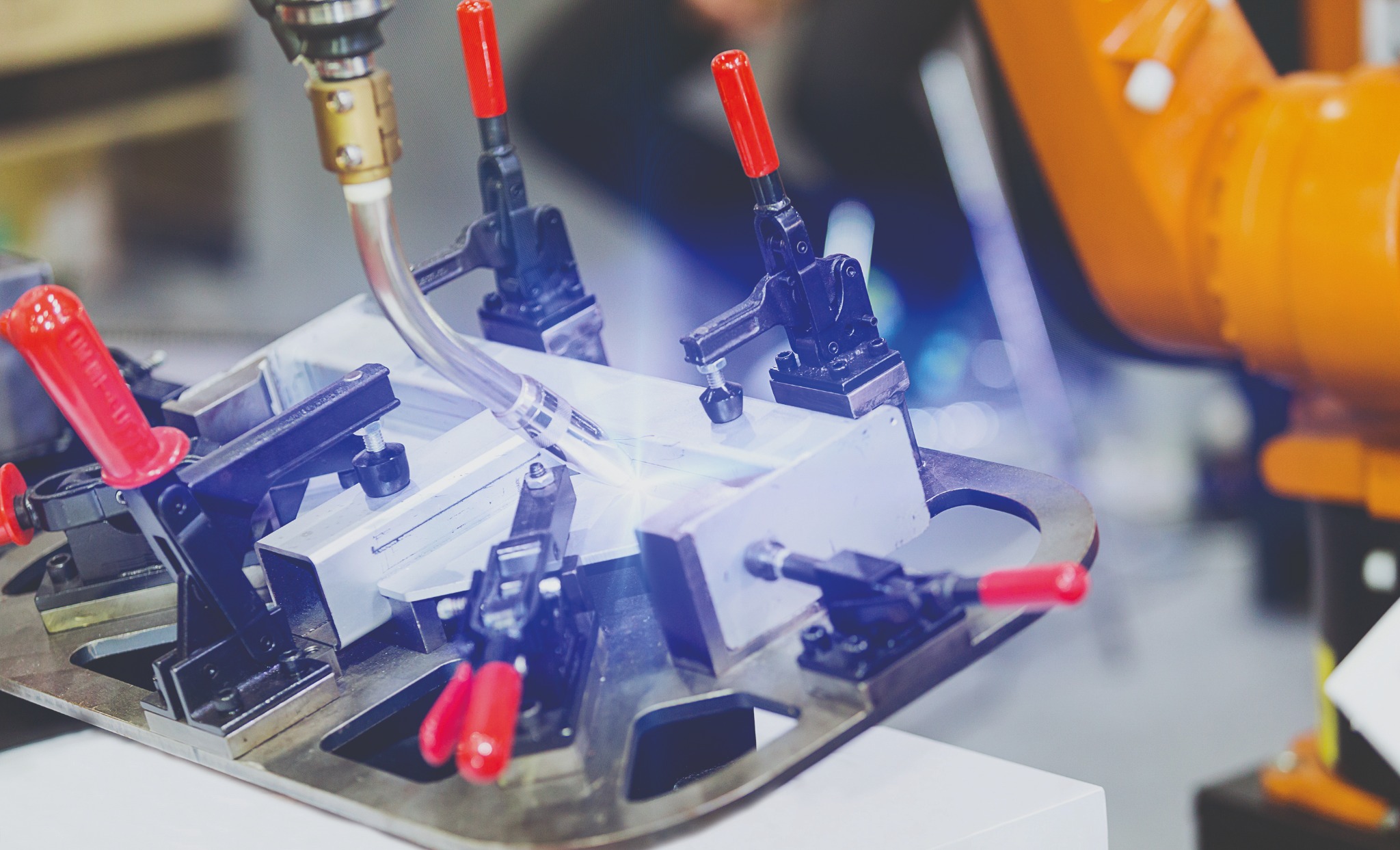 Zrobotyzowane spawanie: pomarańczowy robot spawalniczy w akcji, spawający metalową konstrukcję, z widocznymi mocowaniami z czerwonymi uchwytami.