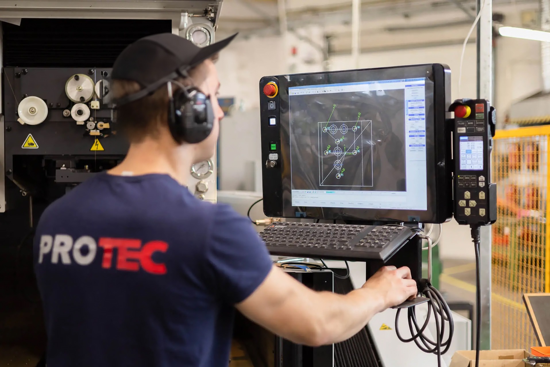 Operator CNC w słuchawkach nadzoruje proces obróbki na maszynie, obserwując schemat na monitorze. Widoczna klawiatura i panel sterowania.