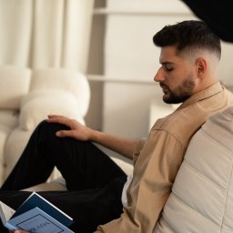 Eryk Basiński - Mężczyzna w beżowej koszuli czyta książkę 'Prada' siedząc na kremowej sofie. Stonowana kolorystyka, miękkie światło, profil twarzy. Sesja w studio.