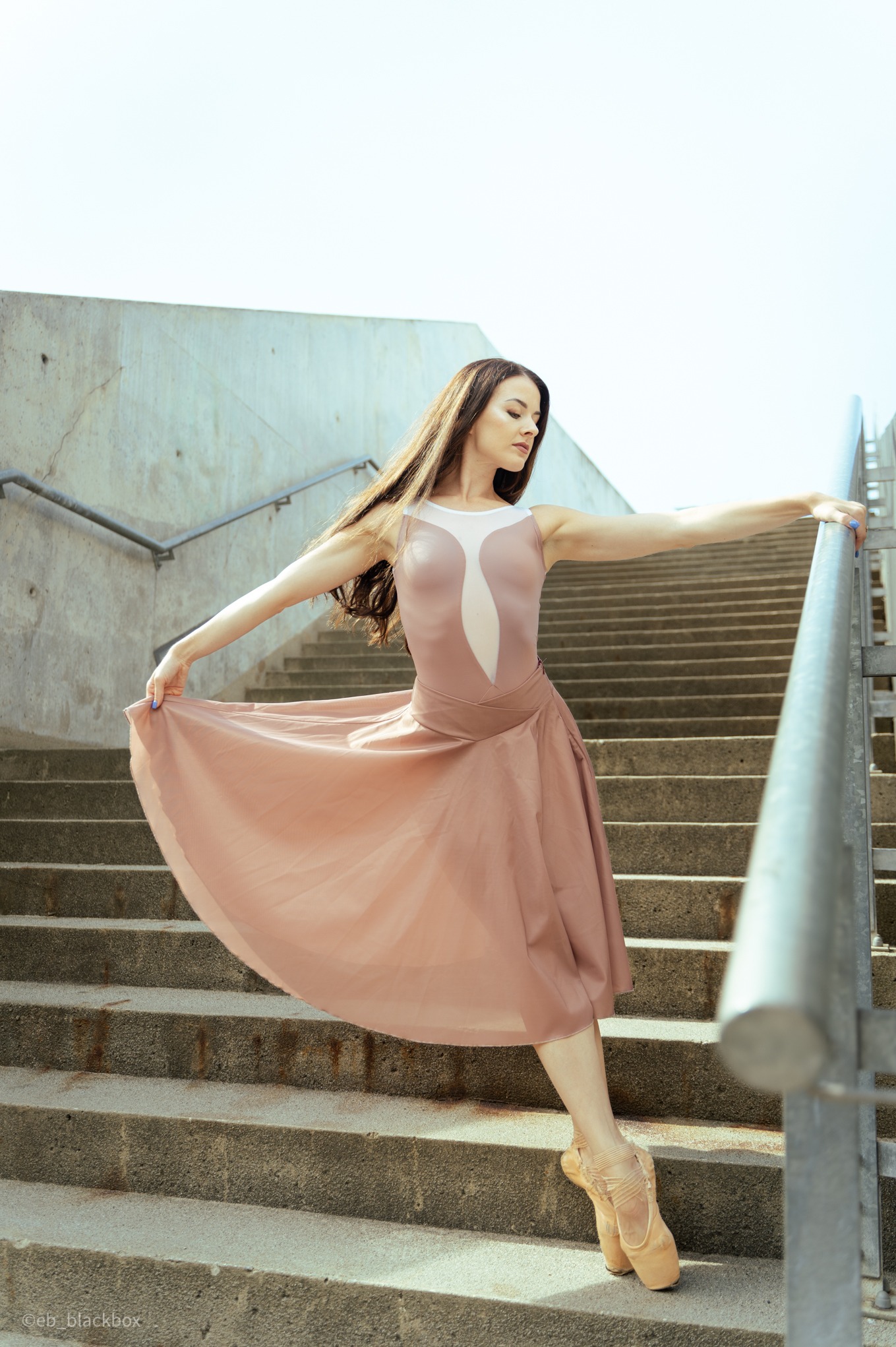 Baletnica w cielistej sukience pozuje na betonowych schodach, trzymając się poręczy. Delikatne światło podkreśla jej długie włosy i taneczną grację.