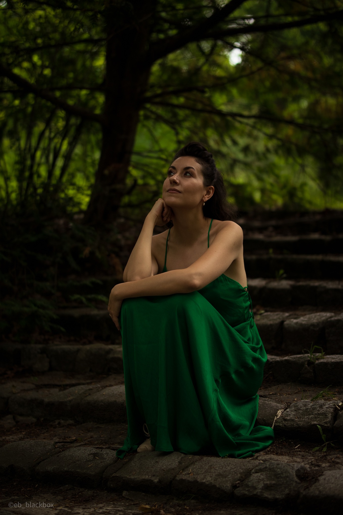 Portret kobiety w szmaragdowej sukni, siedzącej na kamiennych schodach w leśnym otoczeniu, zamyślonej i spoglądającej w górę. Subtelne oświetlenie podkreśla naturalność scenerii.