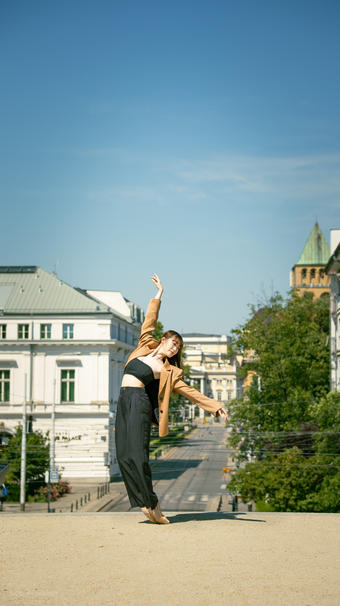 Kobieta w brązowej marynarce i czarnych spodniach tańczy na tle miejskiego krajobrazu z budynkami i błękitnym niebem. Ujęcie z dołu podkreśla dynamikę ruchu.