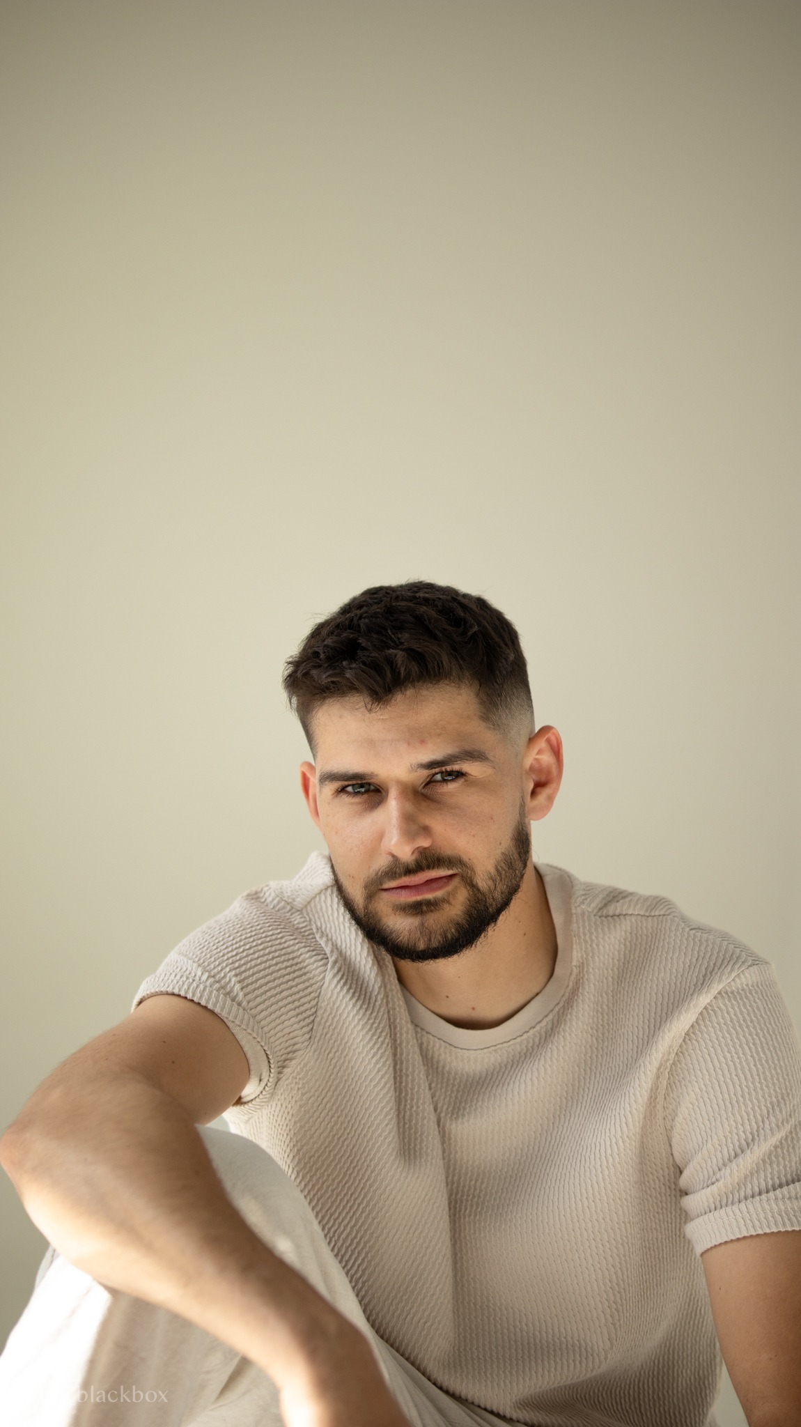 Mężczyzna z brodą w jasnej koszulce, siedzący na podłodze, patrzy w obiektyw. Neutralne tło, naturalne światło. Portret z bliska, kadrowanie od klatki piersiowej w górę.