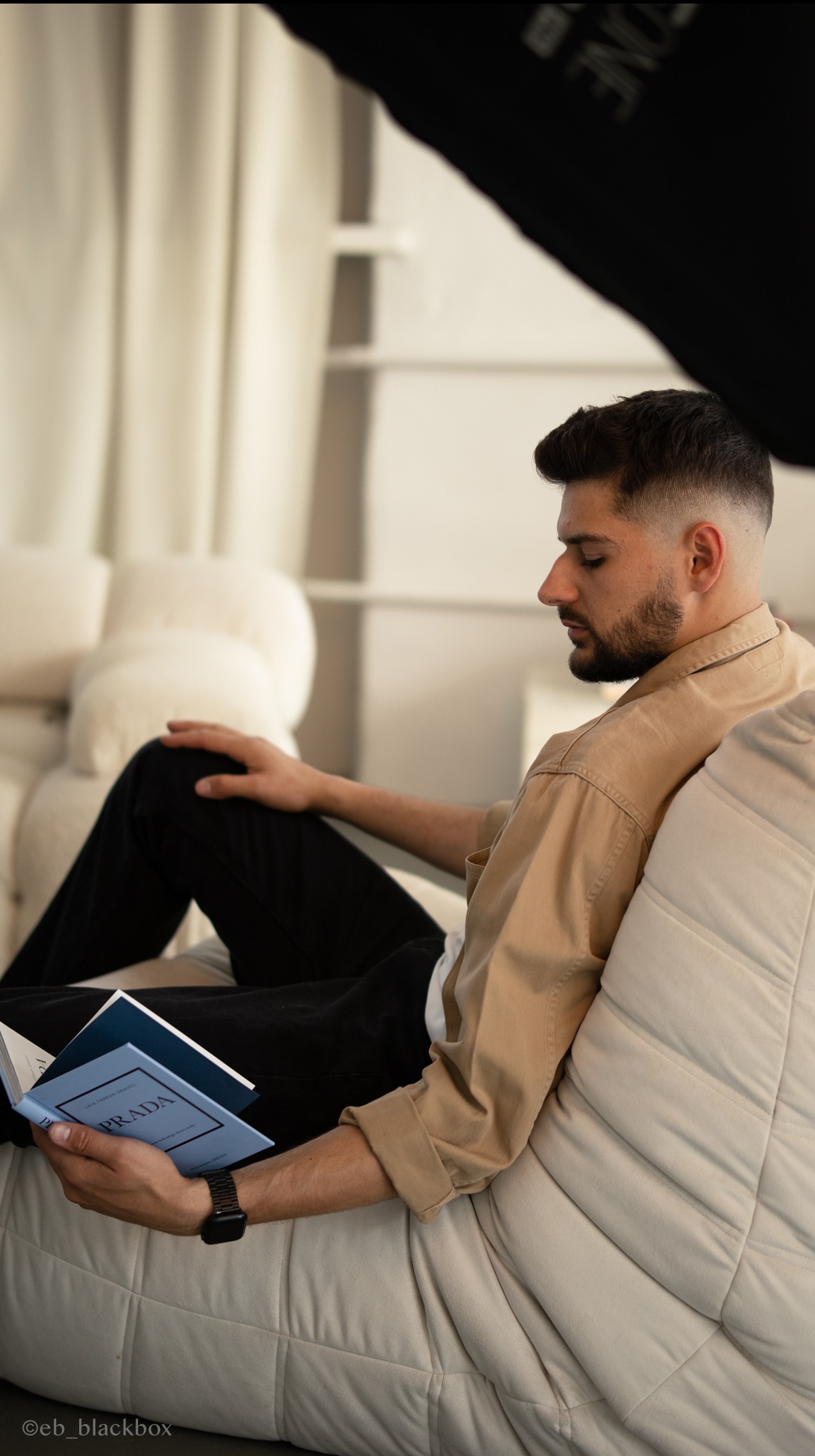 Mężczyzna w beżowej koszuli czyta książkę 'Prada' siedząc na kremowej sofie. Stonowana kolorystyka, miękkie światło, profil twarzy. Sesja w studio.