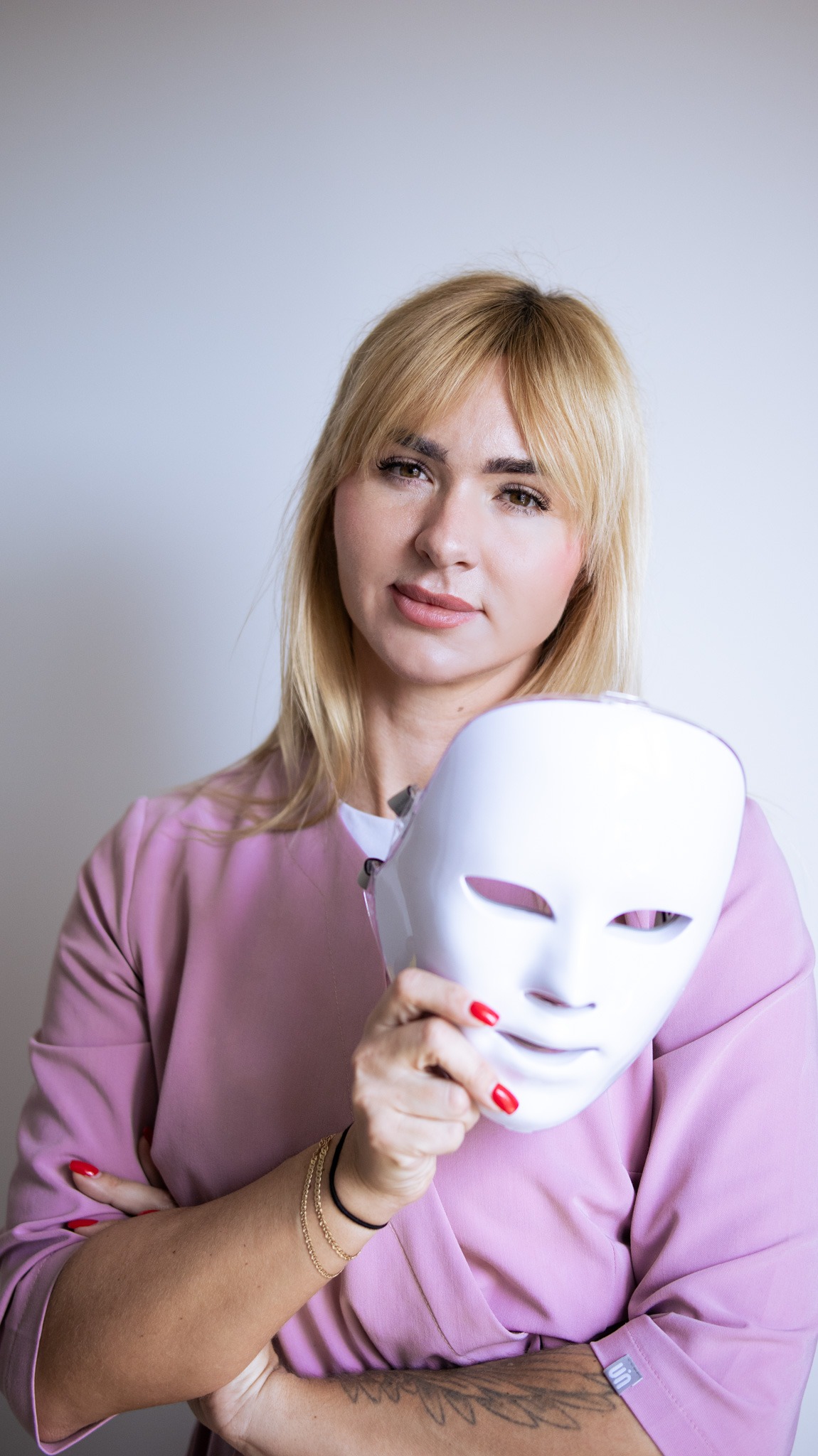 Portret kobiety z blond włosami trzymającej białą maskę LED do fototerapii, w różowym uniformie, z widocznym tatuażem na ramieniu i czerwonym manicure.