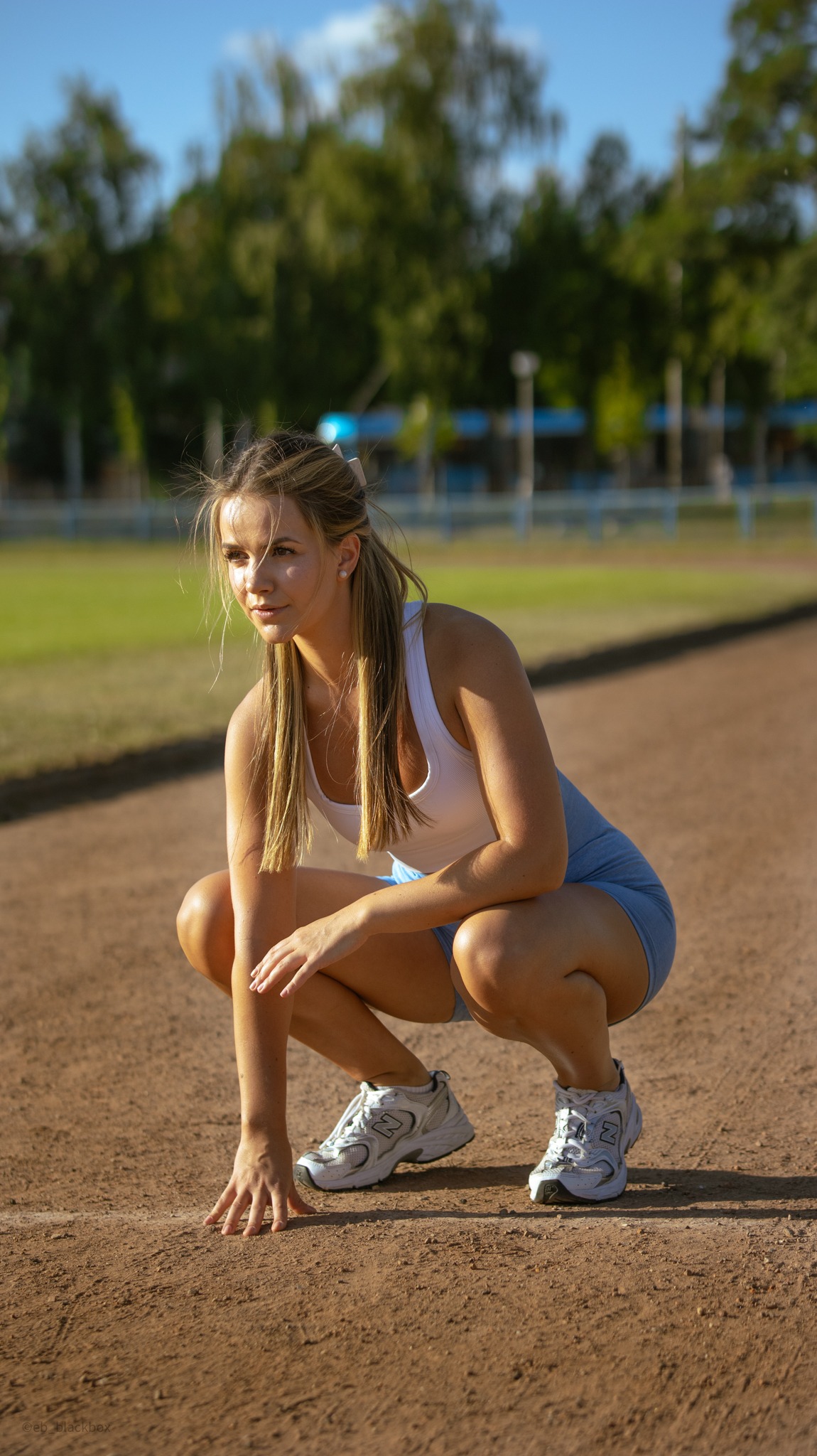 Kobieta w sportowym stroju, w przysiadzie na bieżni, z ręką na linii startu. Gotowa do biegu, skupiona, w tle zieleń i błękit nieba.
