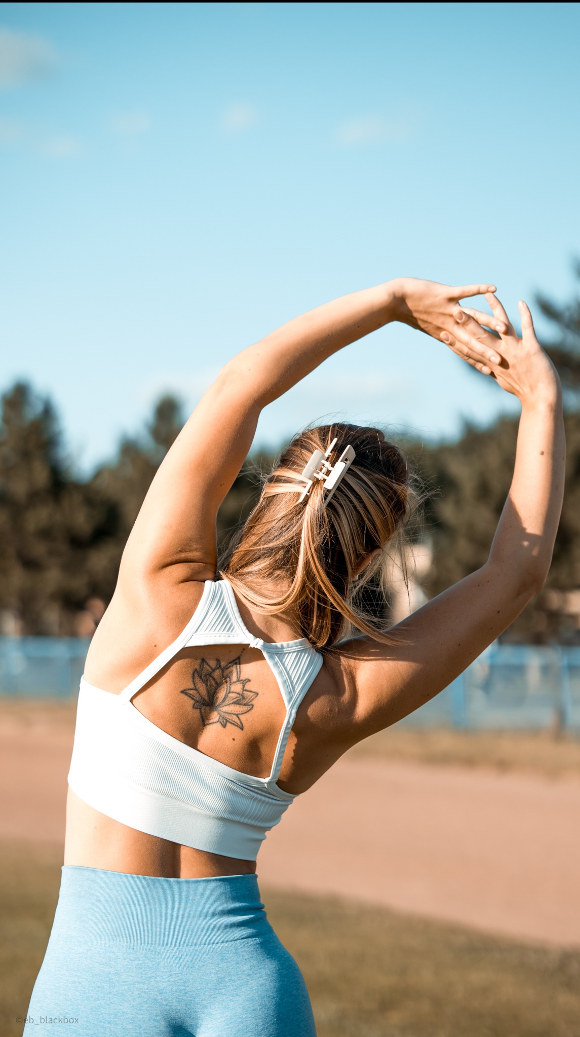 Kobieta w sportowym stroju rozciąga się na tle boiska, widoczny tatuaż lotosu na plecach i spinka we włosach. Dłonie uniesione do góry.