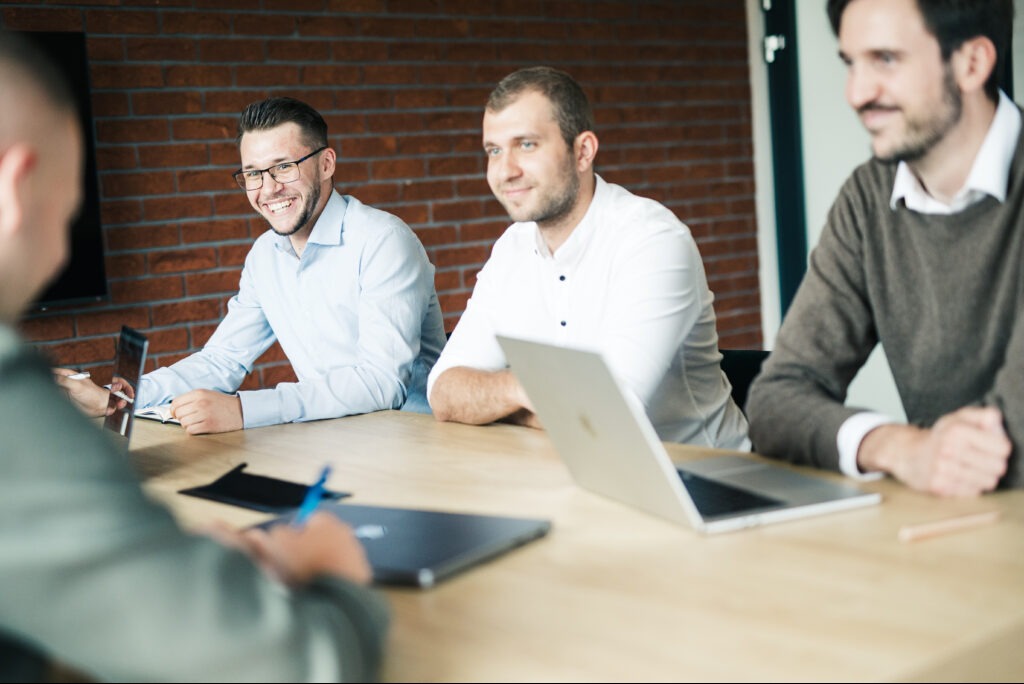 Zespół w biurze podczas spotkania, z uśmiechem i laptopami na stole, ceglana ściana w tle. Jeden z mężczyzn ma okulary.