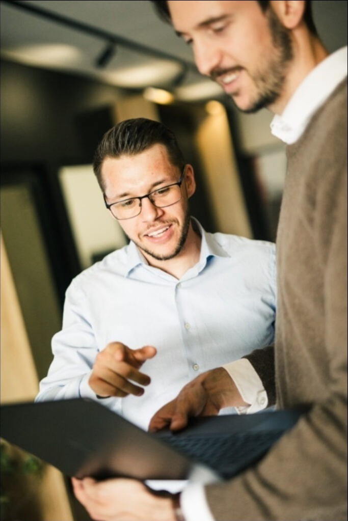 Dwóch mężczyzn analizuje dane na laptopie; jeden z nich wskazuje na ekran, sugerując omówienie strategii lub wyników kampanii reklamowej.