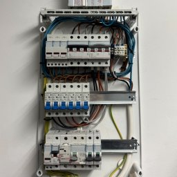 SecVolt Adrian Krupecki - Rozdzielnia elektryczna z zamontowanymi bezpiecznikami i przewodami, widok z przodu. Biała obudowa na jasnej ścianie. Widoczne elementy marki Legrand i Schrack.