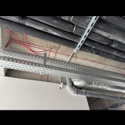 SecVolt Adrian Krupecki - Sufit z widocznymi instalacjami: rury w izolacji, koryta kablowe z przewodami. Szare elementy mocowane do sufitu. Widok z dołu, perspektywa zbieżna.